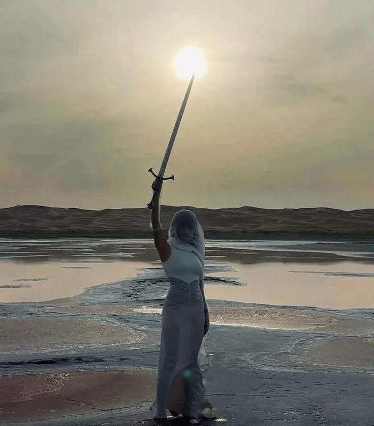 A person dressed as a mermaid standing on a beach holding a sword up towards the sun. The background features a body of water and distant sand dunes or hills. Astrology, moon, spiritual, ritual, ceremony, priestess
