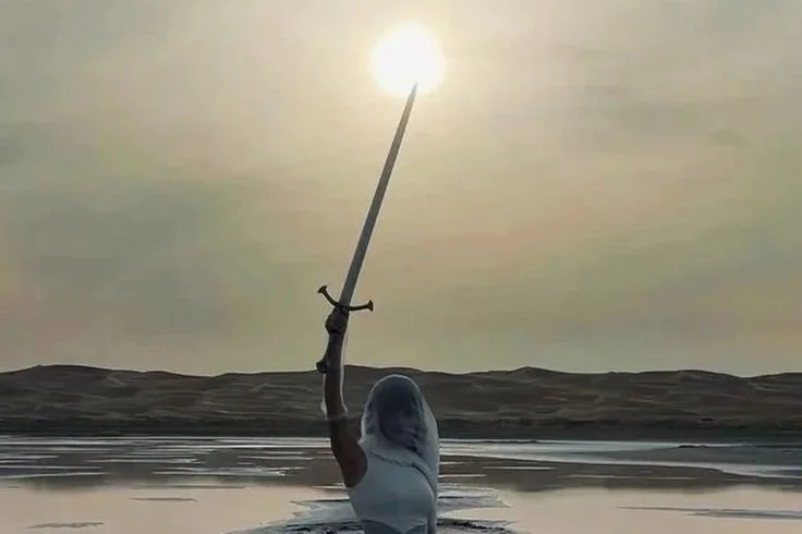 Person in a body of water holding a sword upward, with the sun positioned at the tip of the sword's blade, creating a glowing effect.