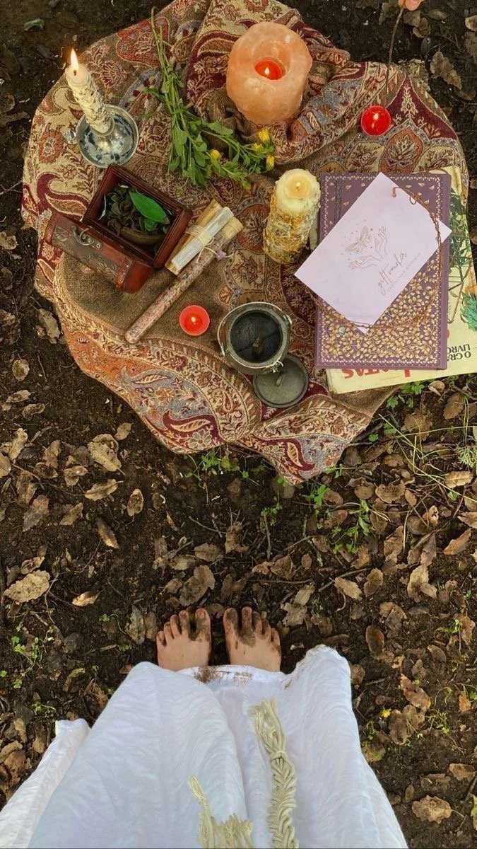A bohemian-style altar setup on a rug outdoors with candles, flowers, and spiritual items, viewed from above with barefoot person in white dress standing on dirt.