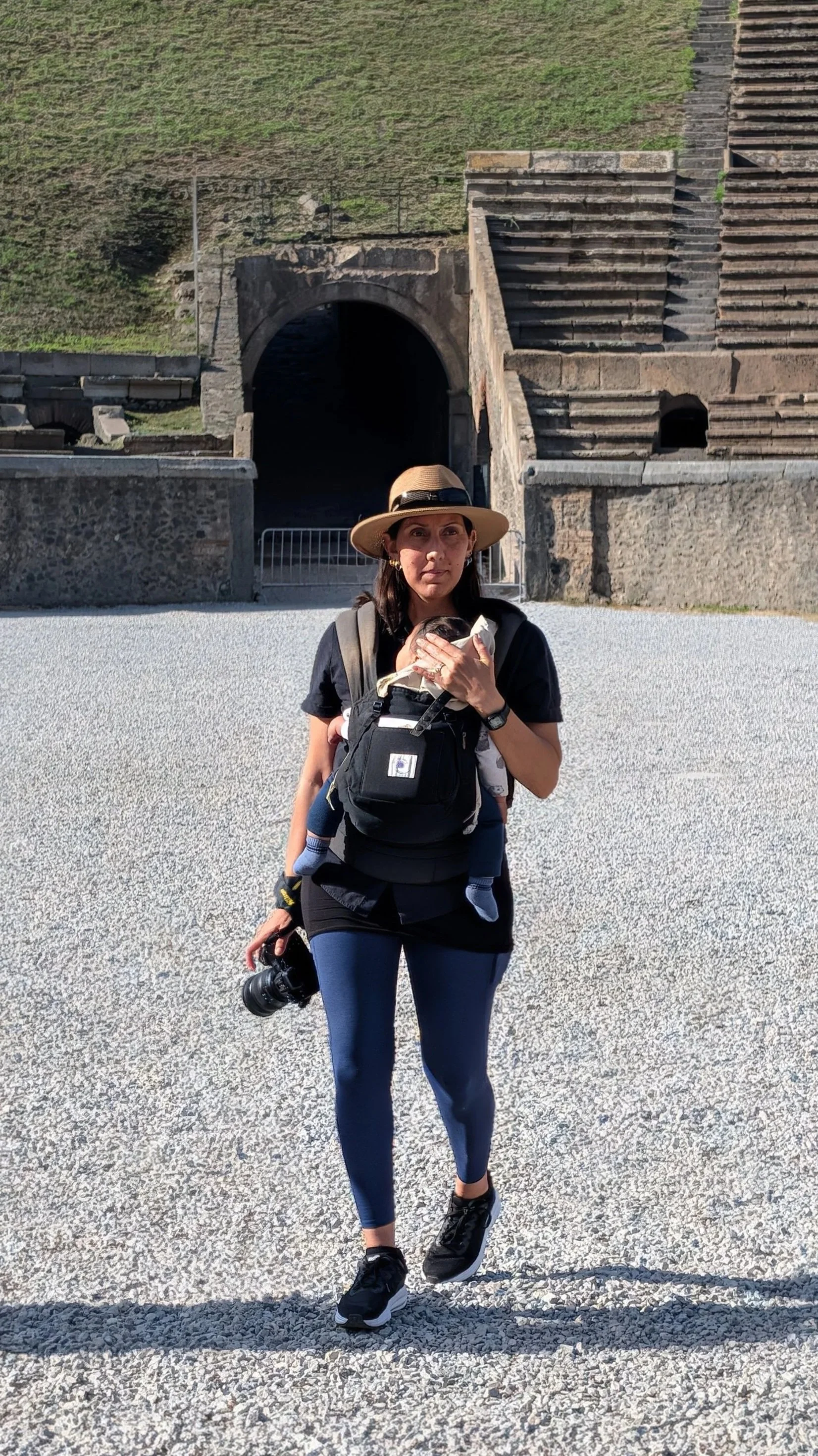 A woman in athletic wear walking outdoors with a camera in her right hand and a child strapped to her chest in a baby carrier. They are in front of a historic stone structure with stairs, tunnel, and grassy hill inside the colosseum in pompeii Italy.