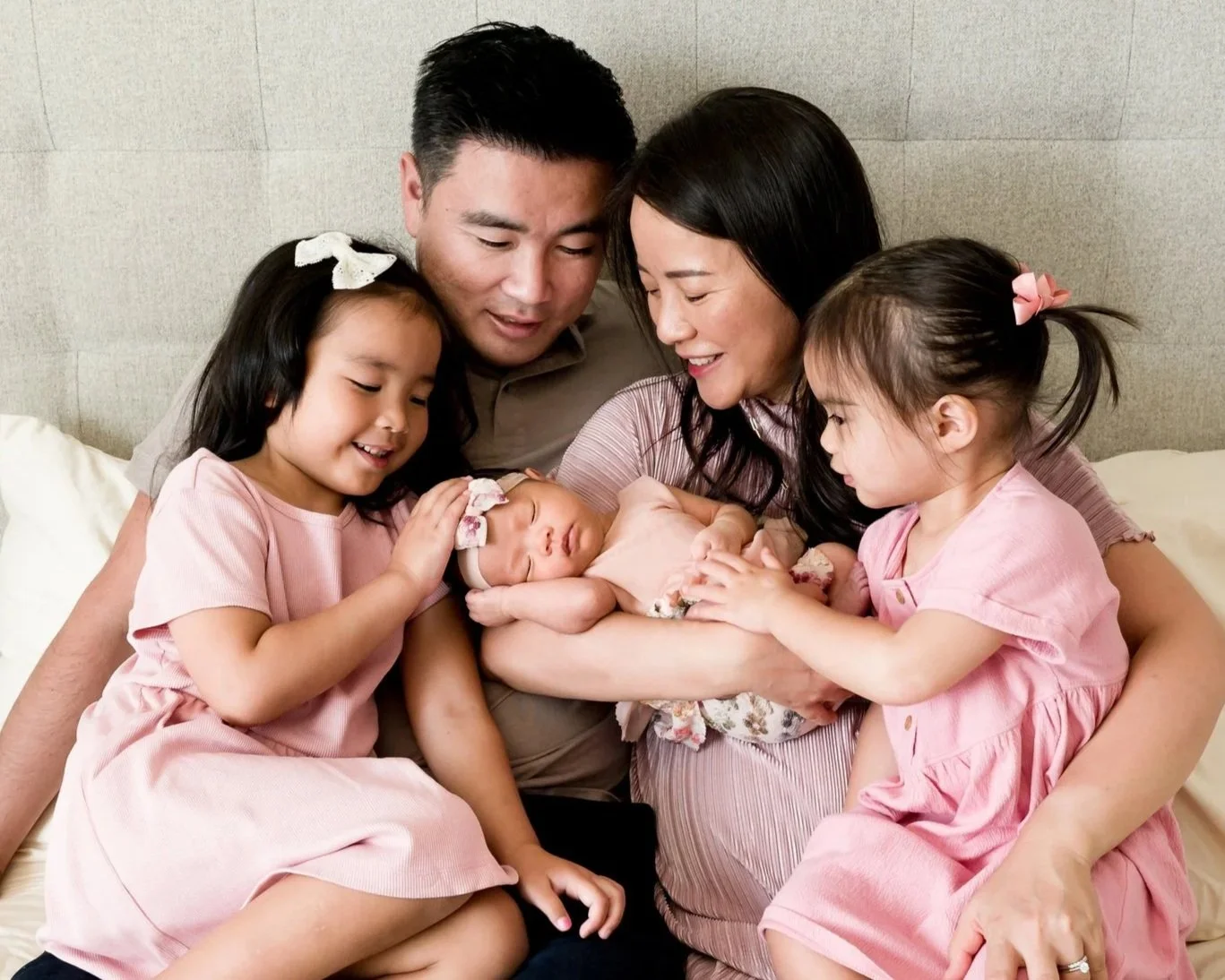 family with 2 little girls and a newborn girl sitting on their bed with a bright and airy natural light. The girls are wearing pink dresses and they are smiling while looking at the newborn baby girl that mom is holding