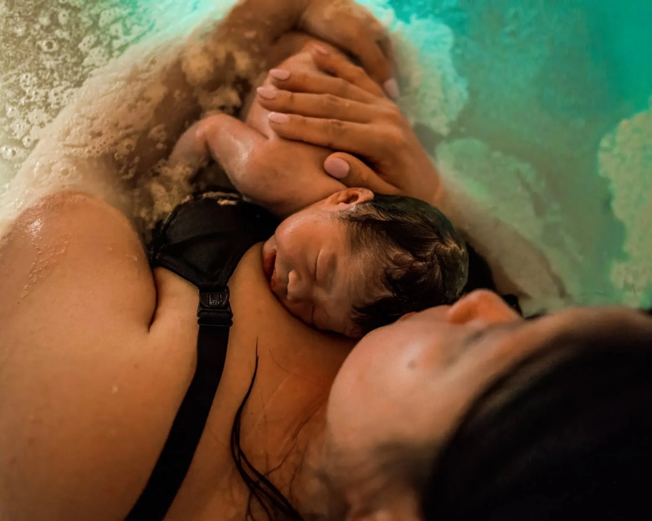 A person with wet hair lying down, holding a baby's head gently, both in water or bath surrounded by bubbles and foam.