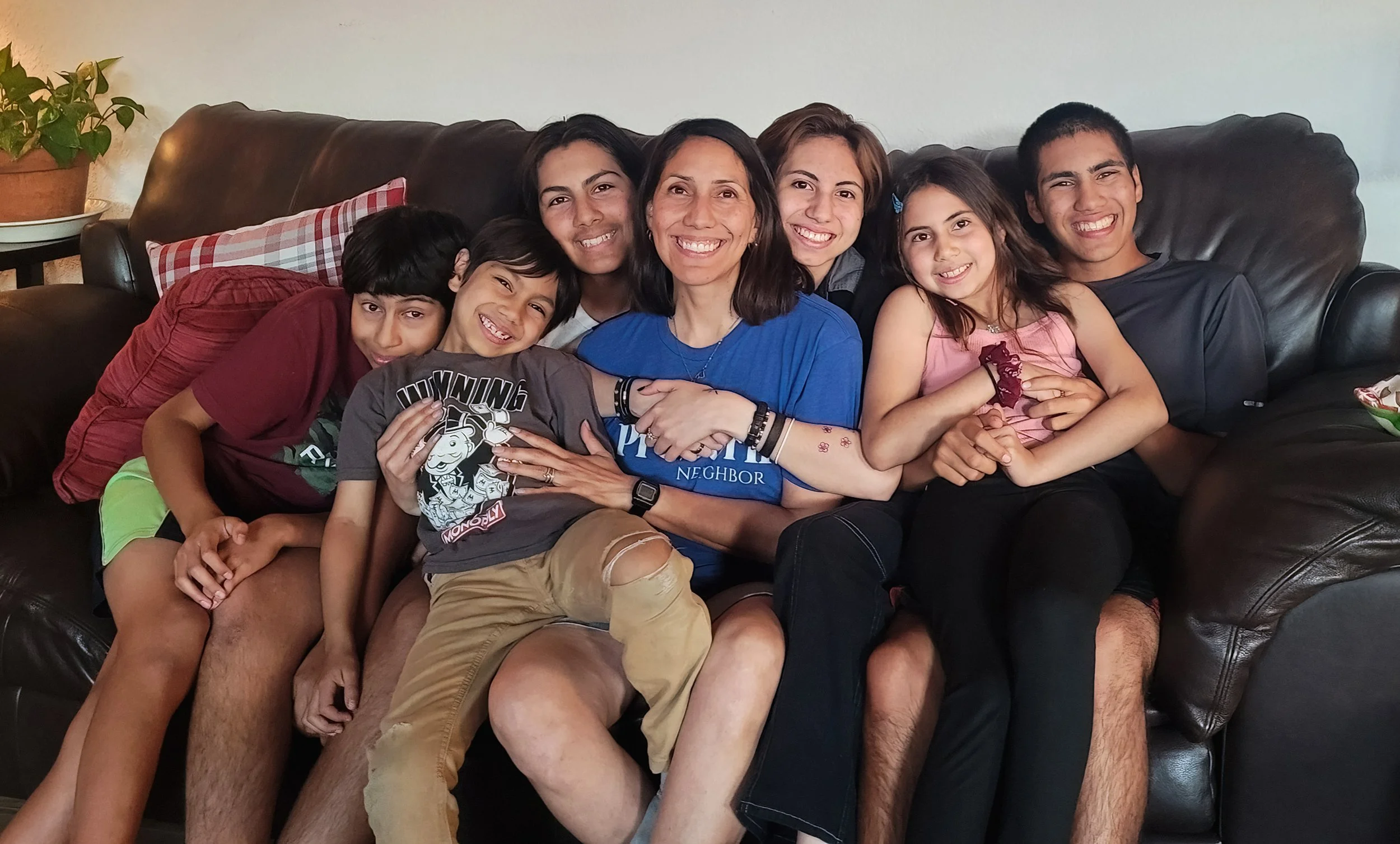 A group of people, including children and adults, sitting closely together on a black leather couch, smiling, in a cozy living room with a potted plant on a side table.