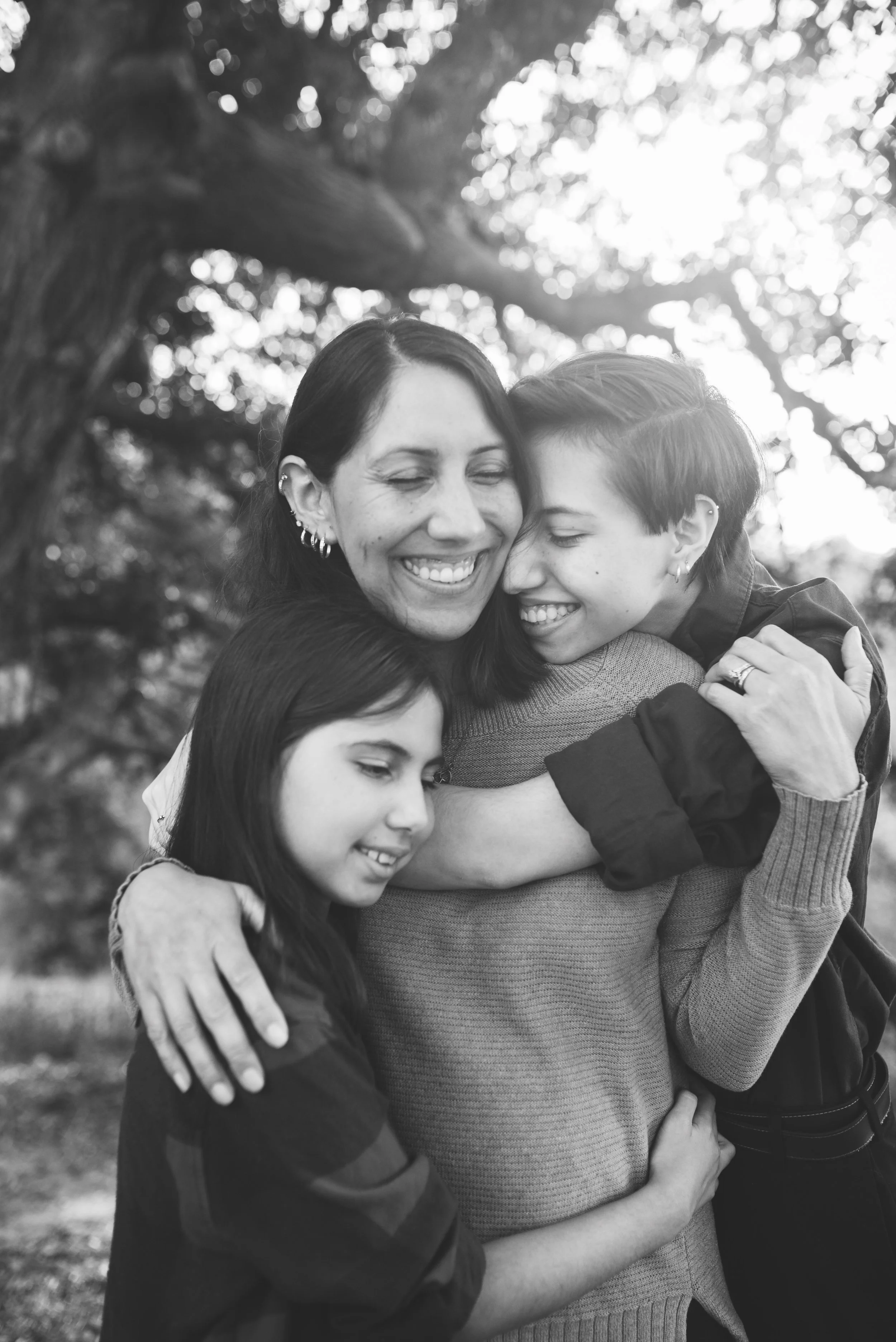 A black-and-white photo of mother and daughters embracing outdoors with trees in the background. They are happily smiling and hugging each other closely.