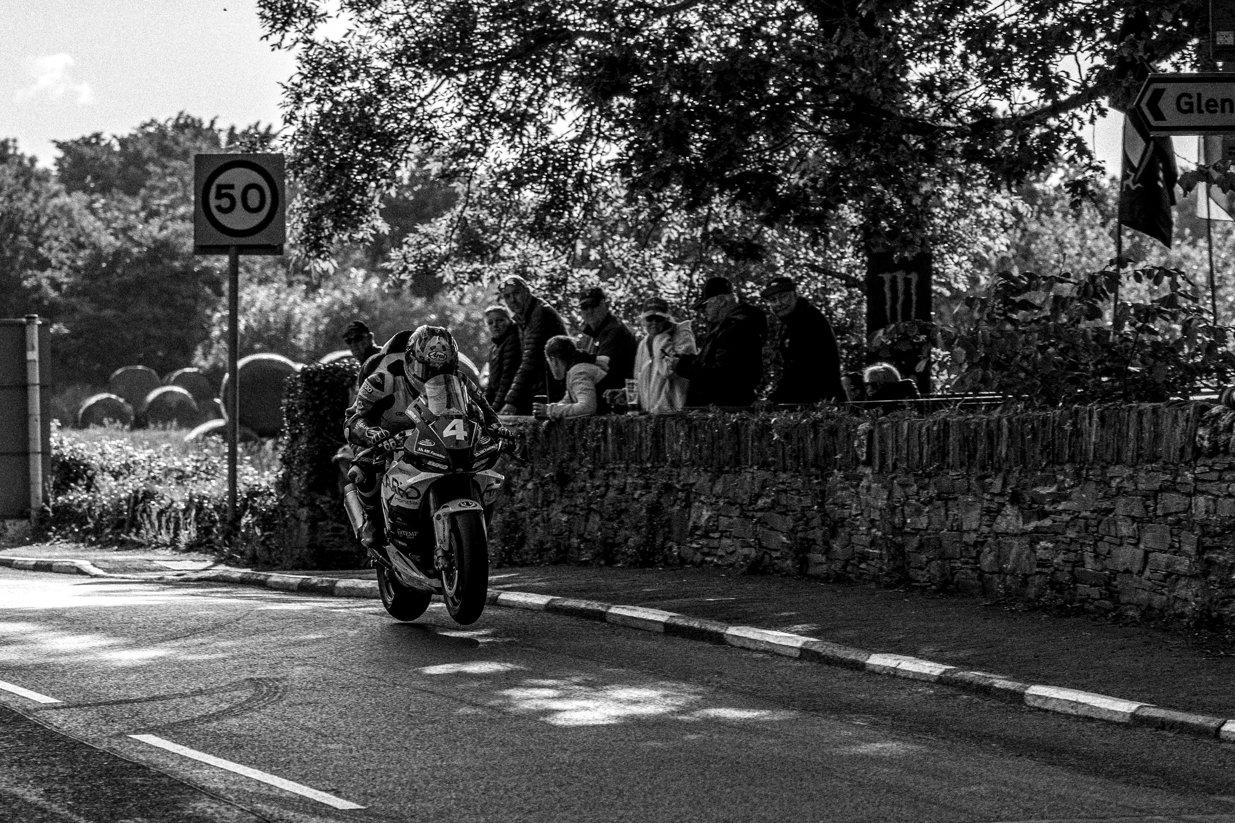 Black and white photo of a motorcycle rider performing a wheelie on a street, with a group of spectators watching behind a stone wall and trees overhead. Traffic signs indicate a 50 km/h speed limit and directions to Gleneagles.