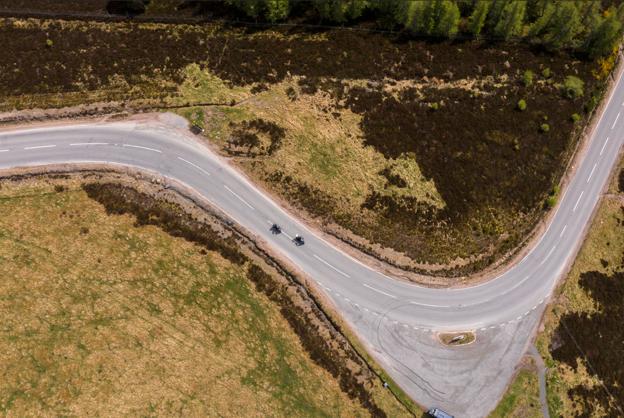 An aerial view of a winding rural road with two motorcycles traveling along it, surrounded by grassy fields and patches of shrubs and small trees.
