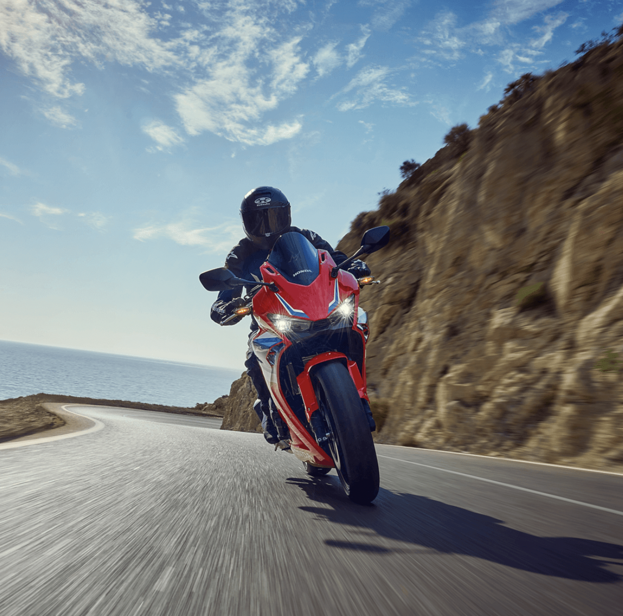 A motorcyclist riding a red and black motorcycle on a curvy coastal road with cliffs and the ocean in the background.