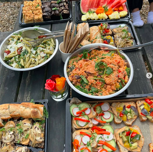 Assorted picnic food on a black wooden table, including pasta salad, vegetable wraps, fruit slices, and open-faced sandwiches with vegetables and spread.
