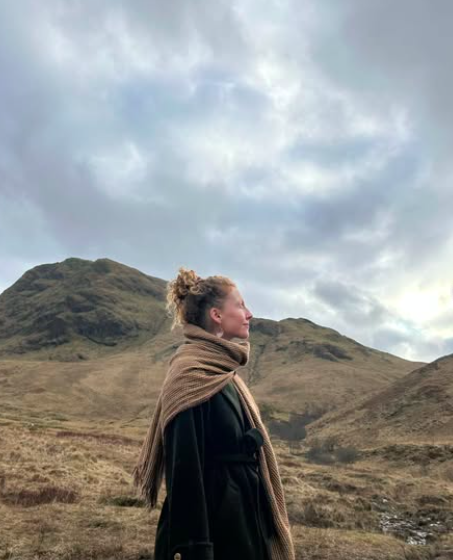 A woman standing outdoors on a grassy hillside with rolling hills and cloudy sky in the background.