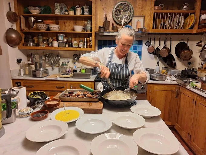 A woman cooking in a kitchen with wooden cabinets, preparing food in a frying pan with several empty plates arranged on the counter in front of her.