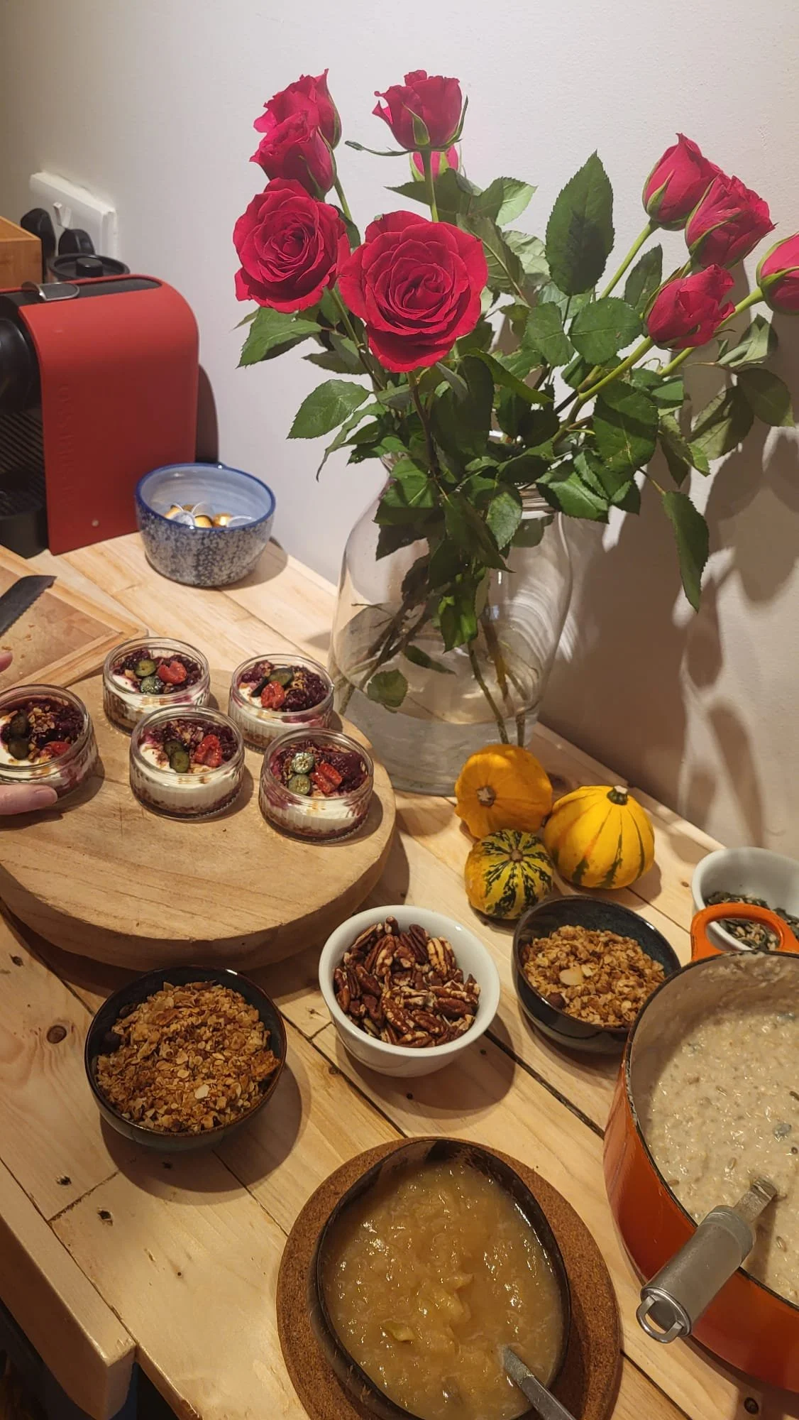 A wooden table with several bowls of food, including granola, pecans, and what appears to be oatmeal or porridge. A round wooden tray holding small dessert jars topped with berries and seeds is also on the table. Behind, there is a bouquet of red roses in a clear glass vase and some small decorative gourds. To the side, a red appliance and a blue bowl with wrapped candies are visible.