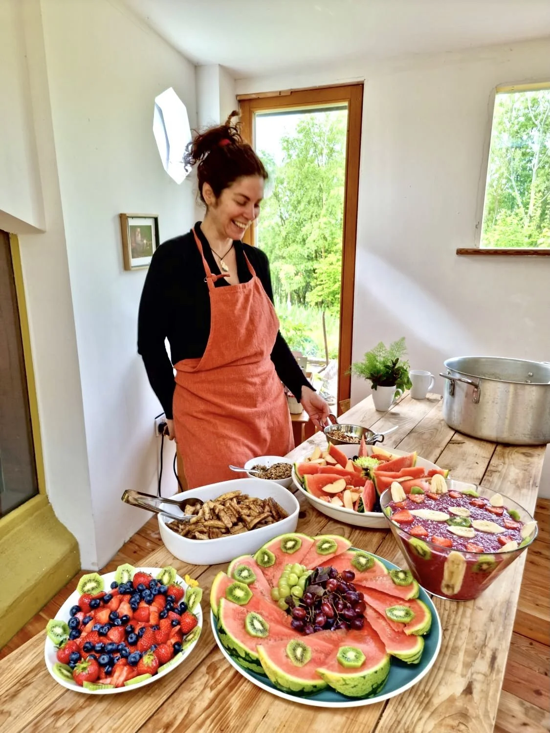 Woman in a coral apron preparing a variety of fresh fruit in a bright kitchen with large windows and natural light.
