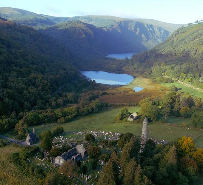 A rural landscape with rolling green hills, two lakes, and a small church or old stone building in the foreground, surrounded by a cemetery and trees.