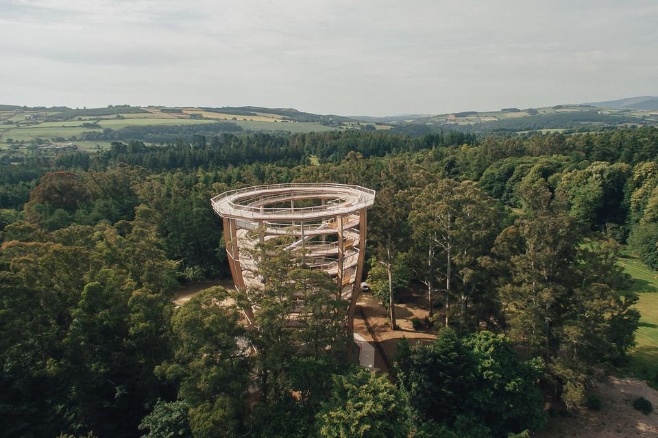 A tall, modern observation tower surrounded by dense green trees and countryside landscape with rolling hills in the distance.