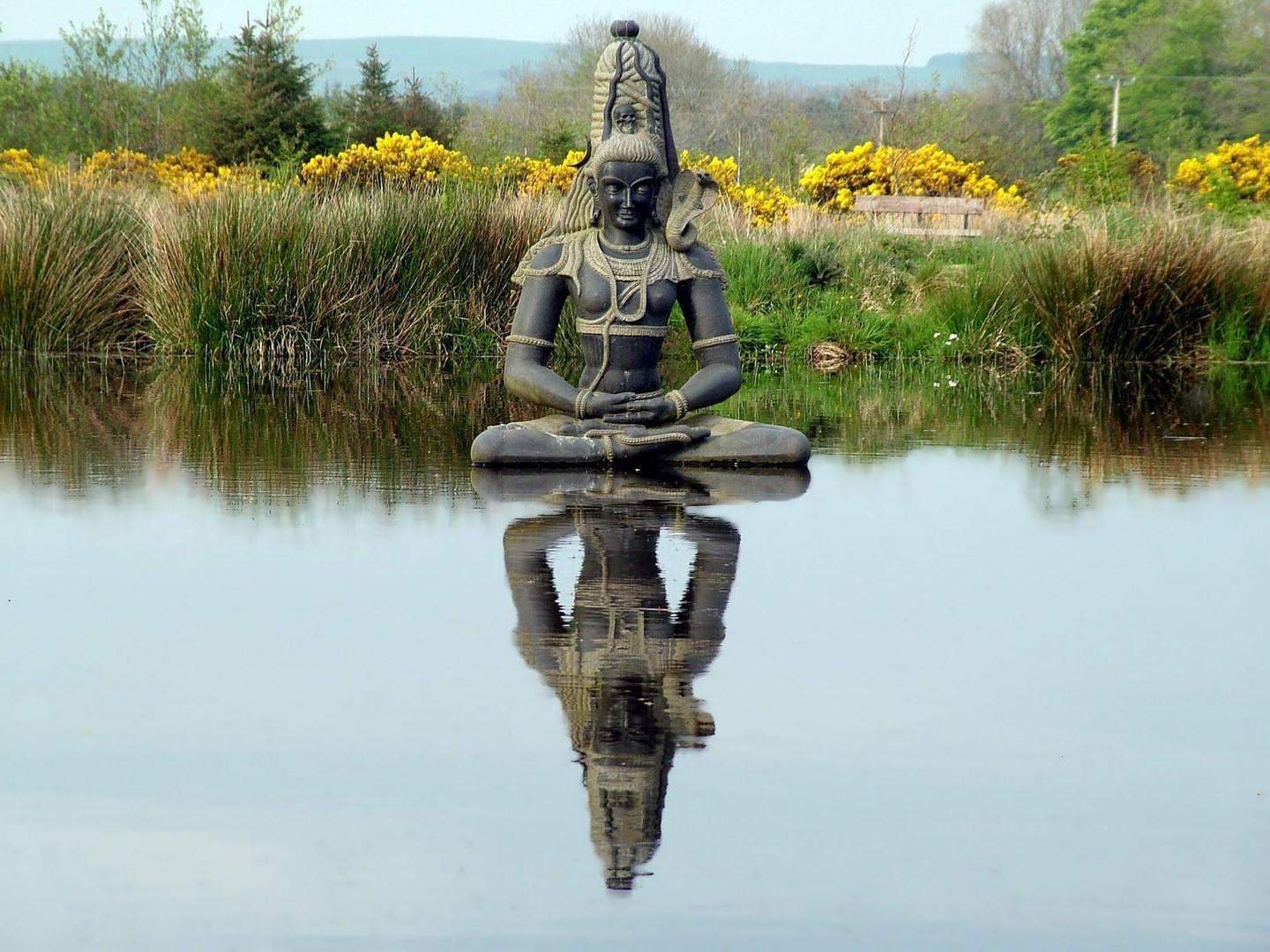 A large black sculpture of a seated Hindu deity with multiple arms is in a pond, with its reflection visible in the water. The background features yellow flowers, trees, and a partly cloudy sky.