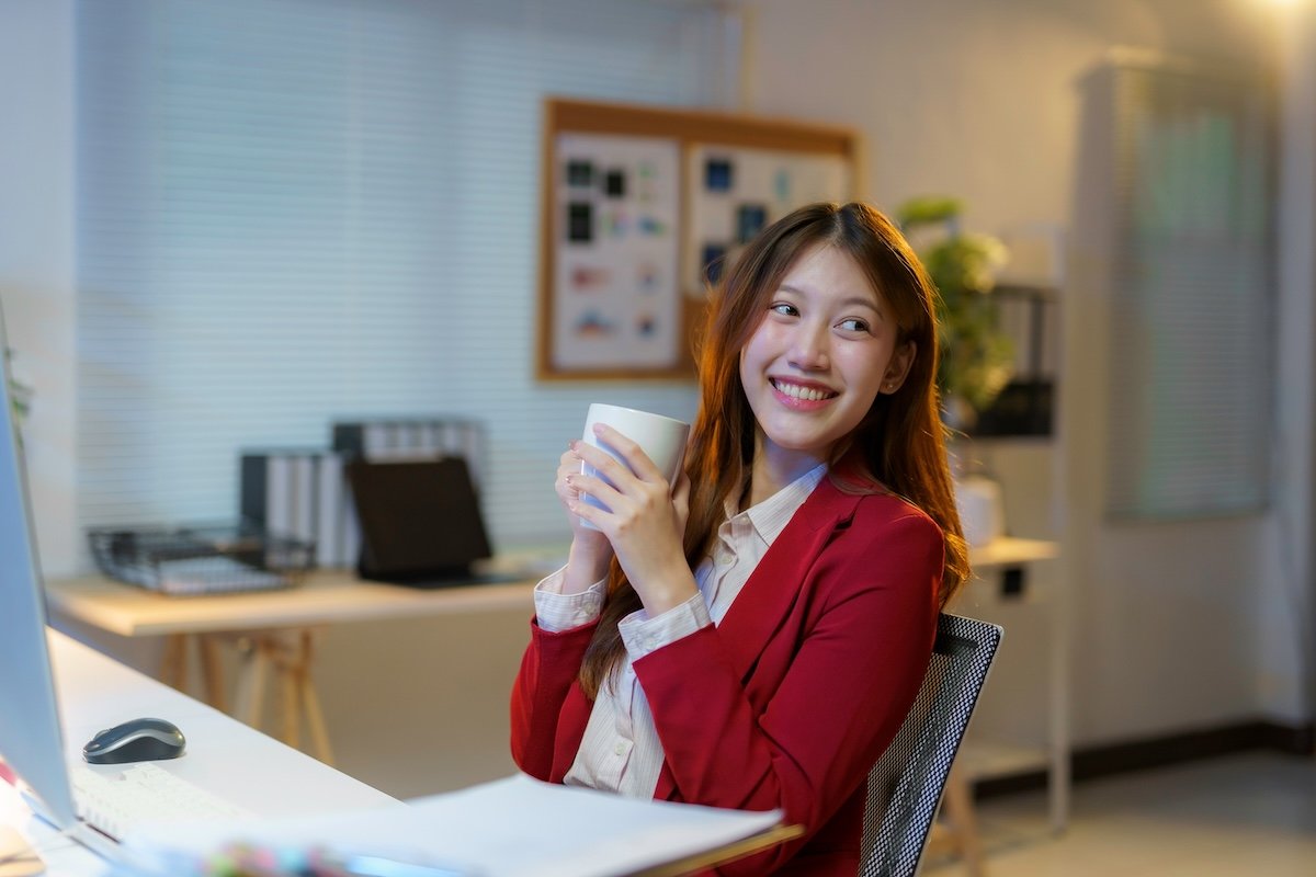 A young woman in a red blazer smiling and holding a white mug in an office environment with a desk and computer.