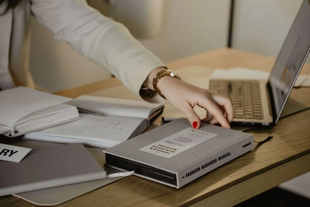 Fashion designer at a laptop reviewing EPR compliance requirements with The Fashion Business Manual and garment sketches
