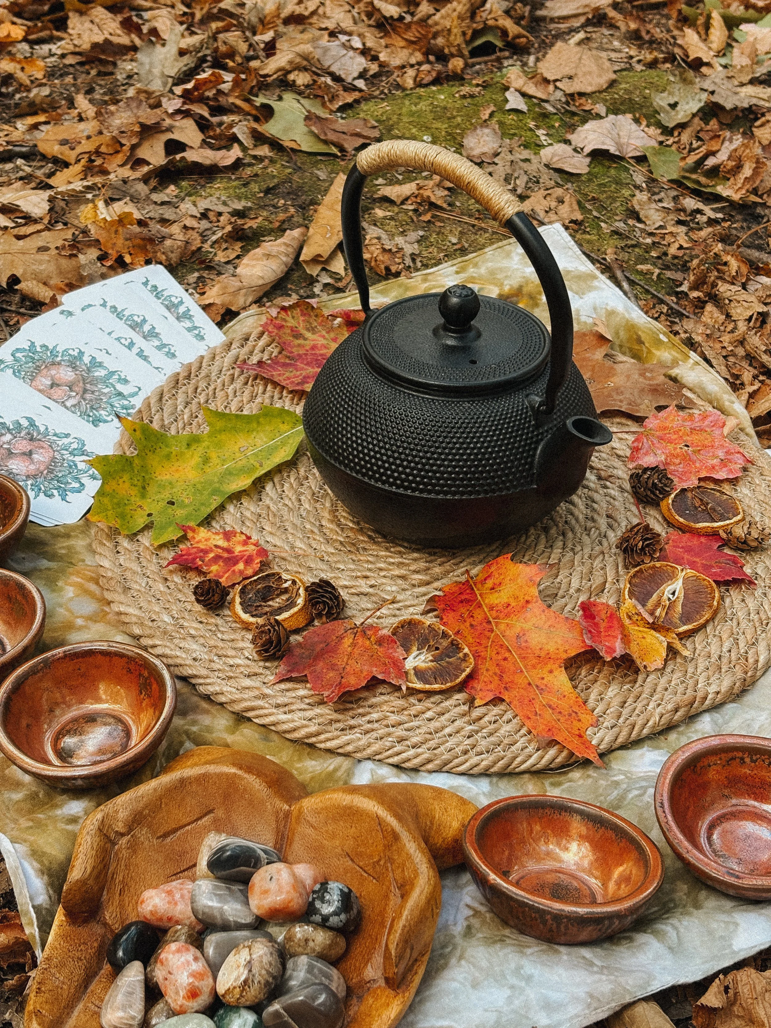A black cast iron teapot on a woven placemat surrounded by autumn leaves, pinecones, dried citrus slices, small ceramic bowls, and decorative stones on a forest floor.