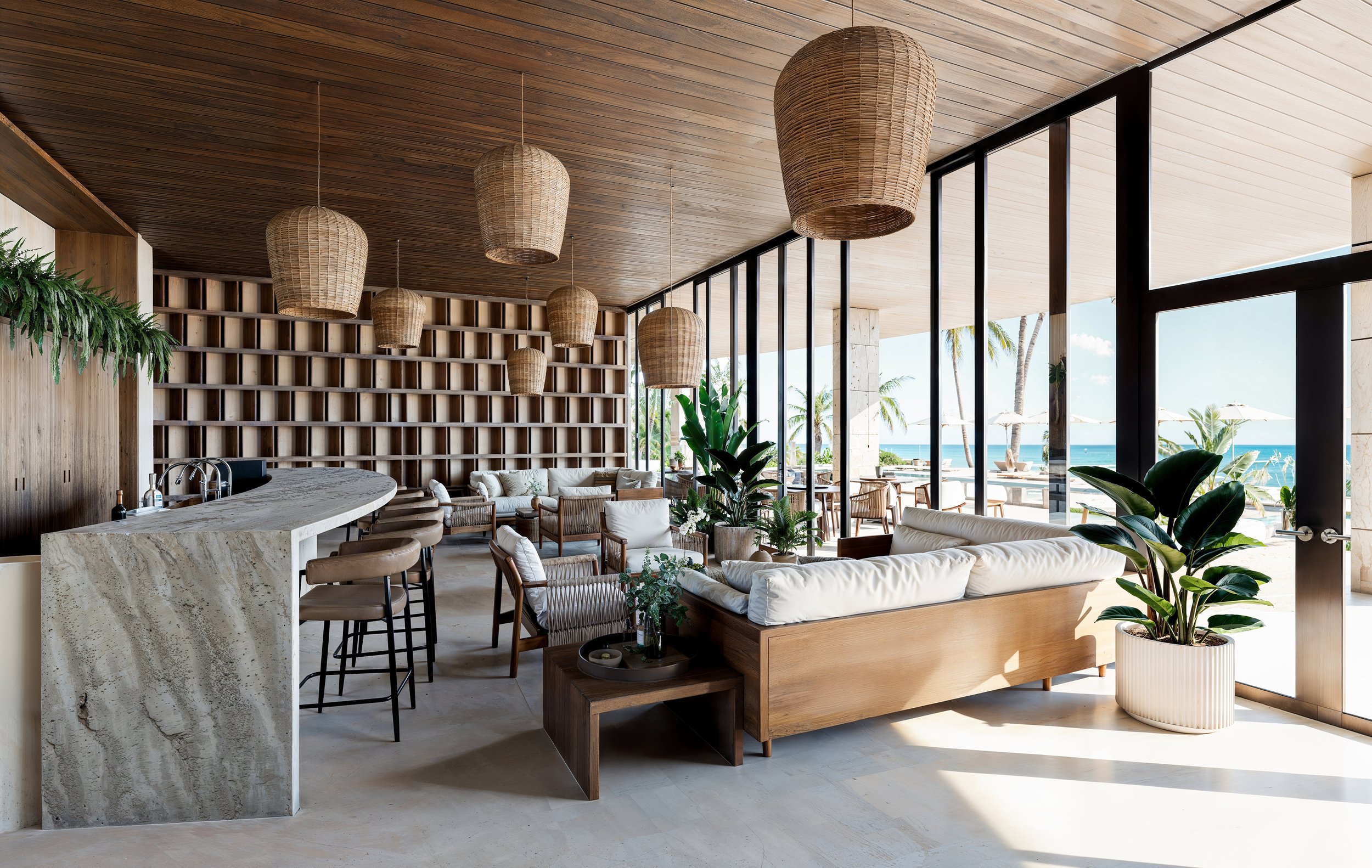 Interior de un salón con muebles de madera, sillones blancos y plantas verdes, con vista al mar y terraza con sombrillas afuera.