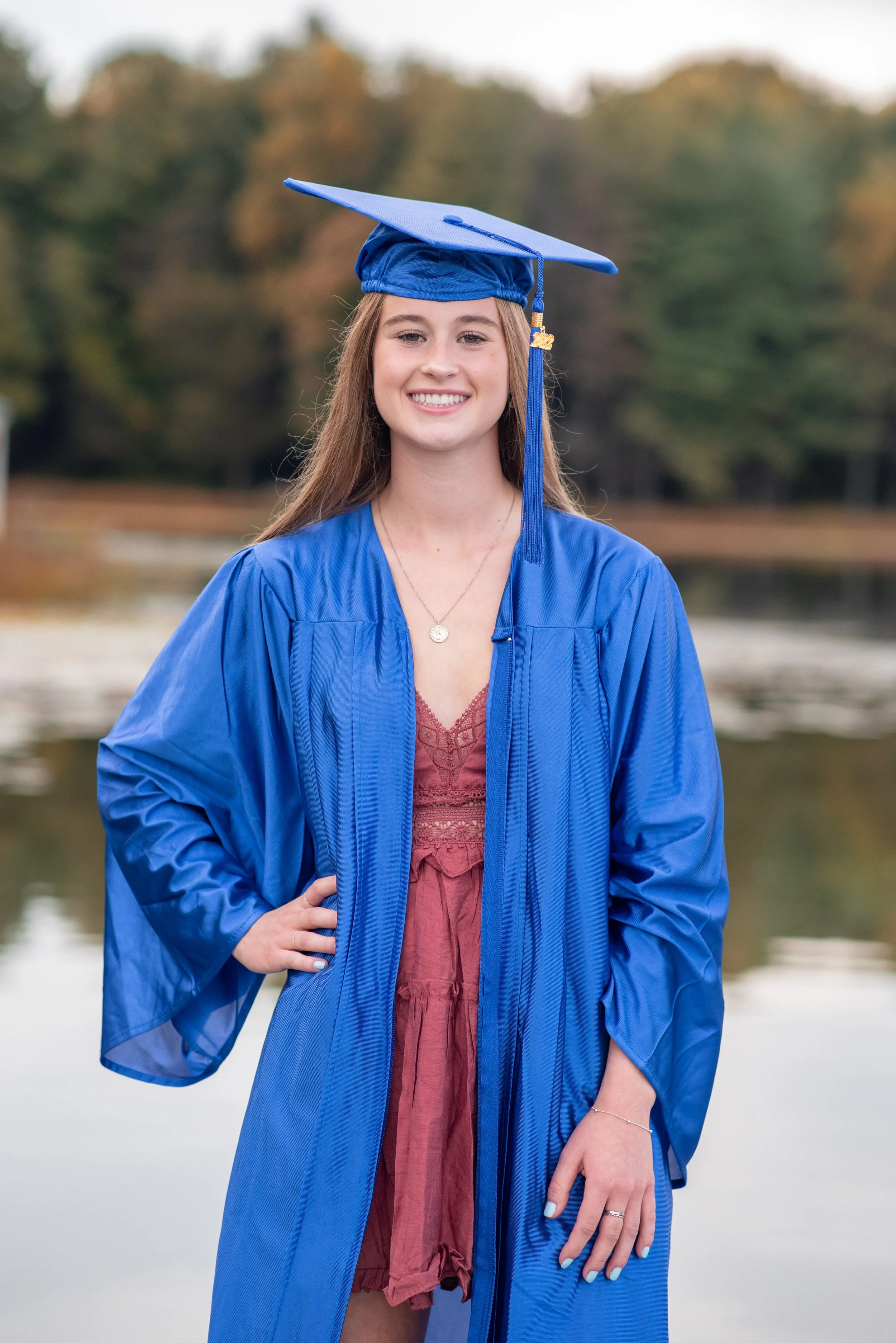 A young woman in a blue graduation gown and cap stands outdoors near a body of water, smiling at the camera. The background features trees with autumn foliage.