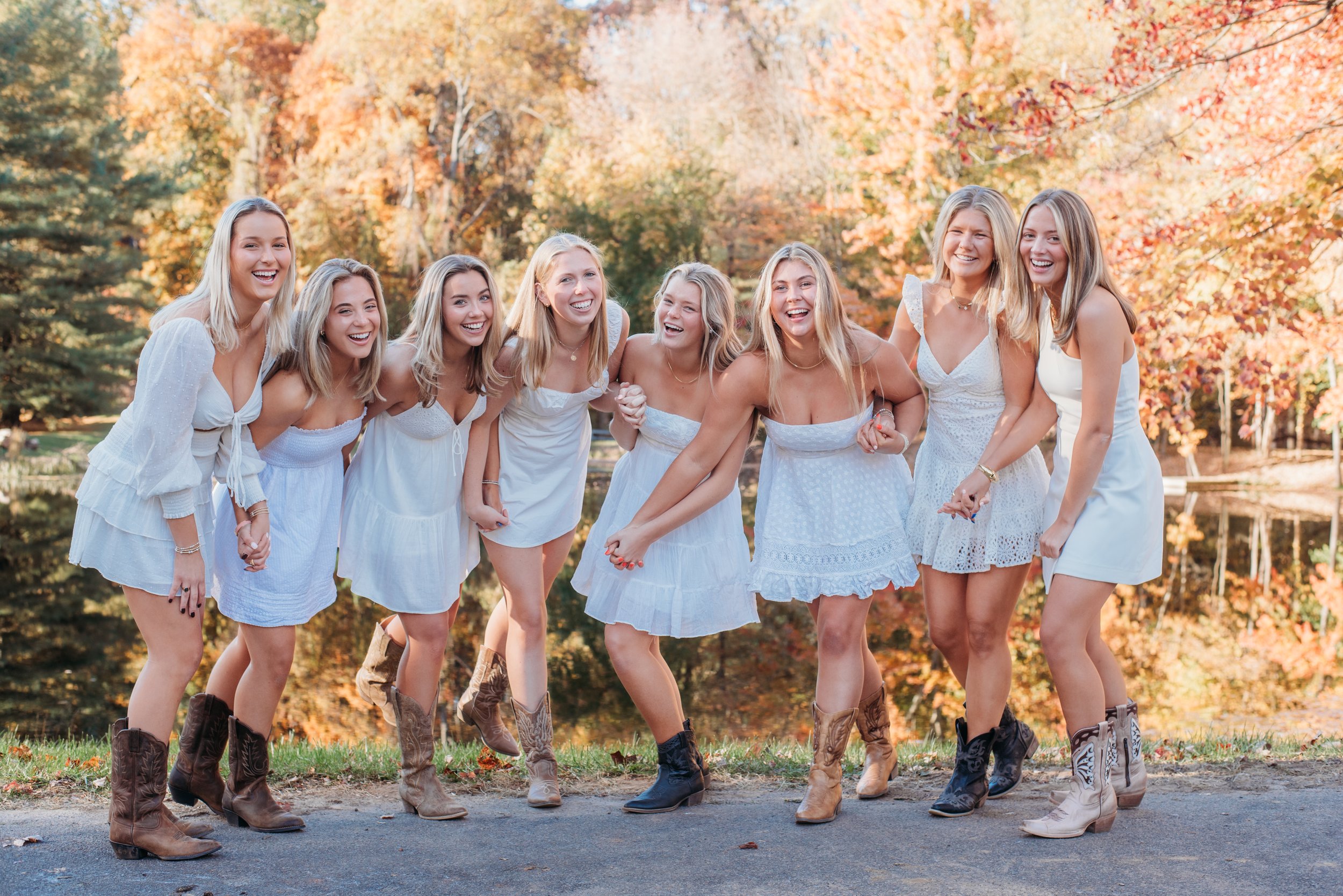 A group of nine young women in white dresses and cowboy boots standing together outdoors in autumn, smiling and enjoying each other's company.