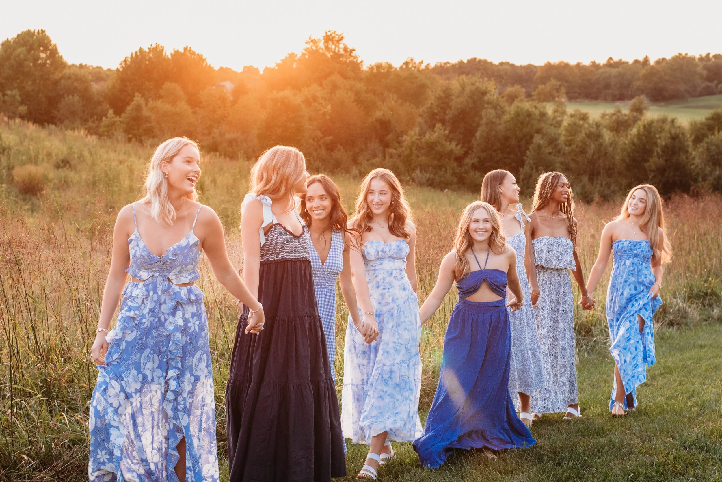 A group of nine women wearing dresses holding hands and walking through a grassy field during sunset, smiling and enjoying each other's company.