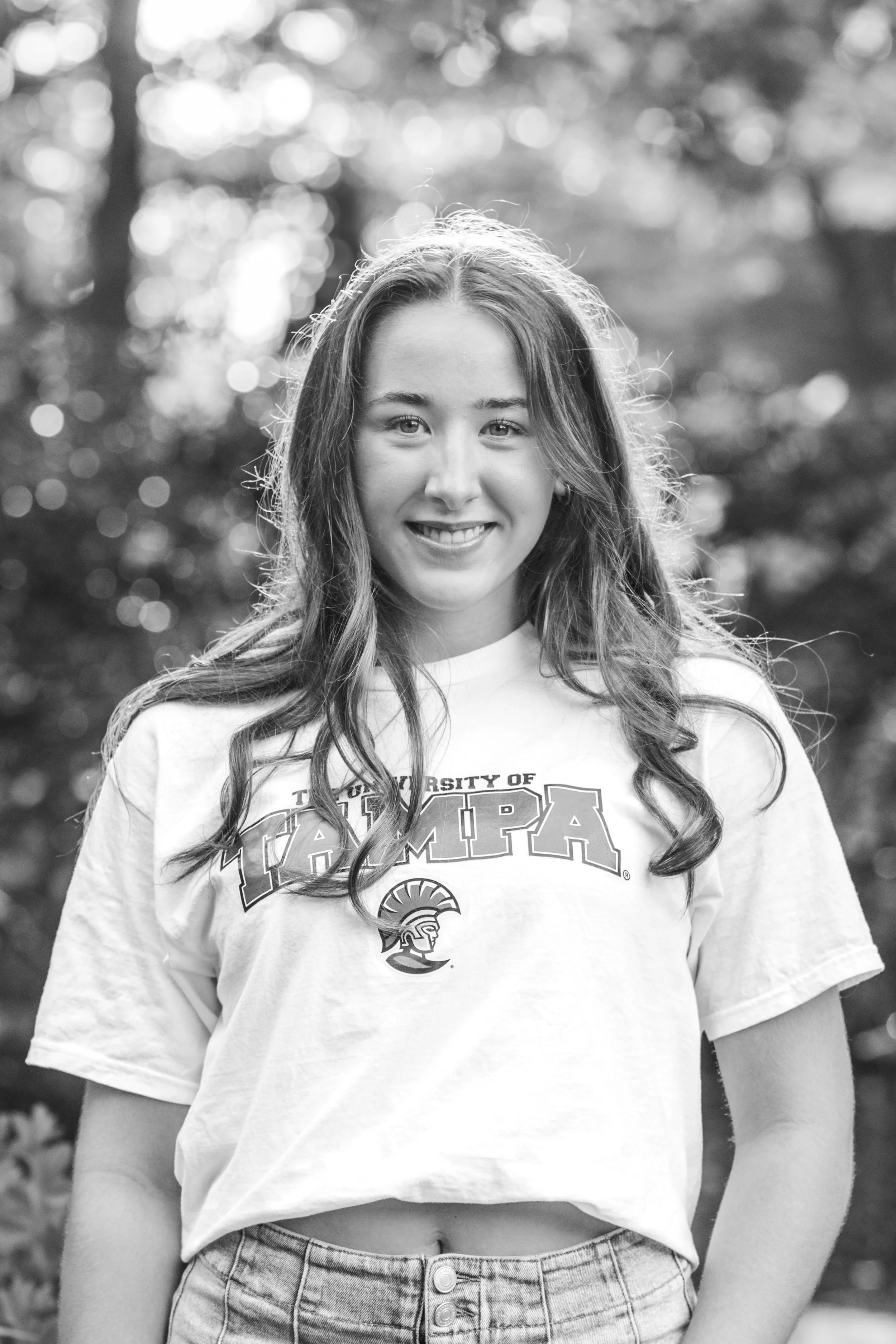 Black and white photo of a young woman smiling outdoors, wearing a Tampa shirt and jeans.