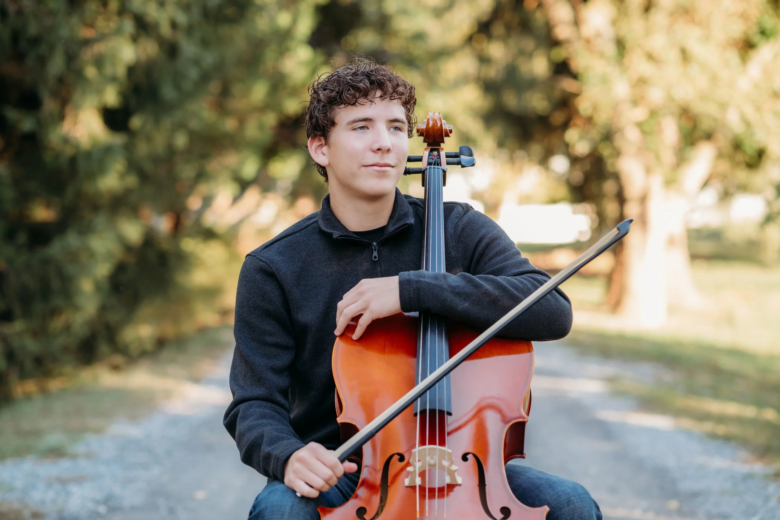 Teen boy playing cello outdoors in a park with trees and sunlight.