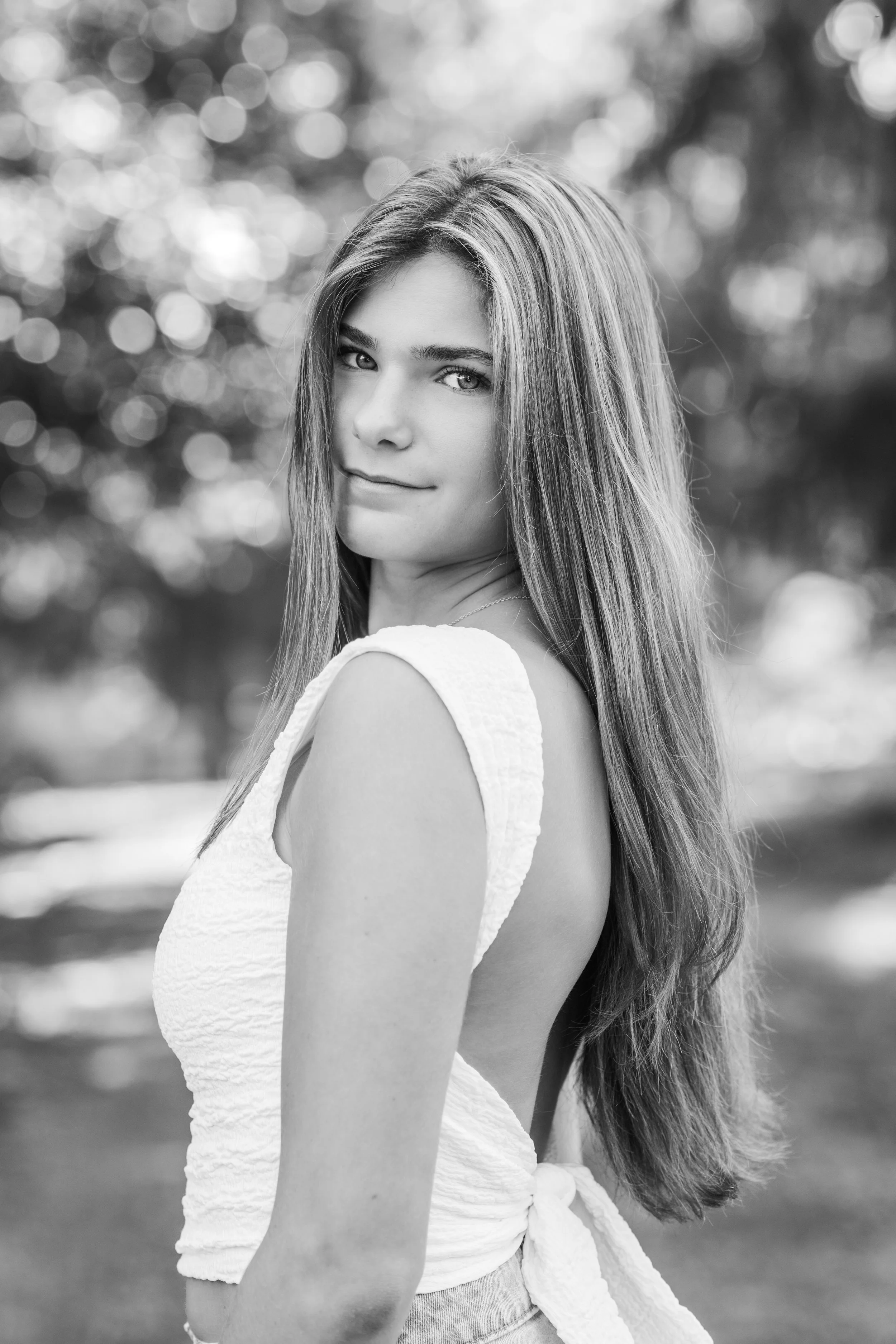 Black and white portrait of a woman with long hair outdoors, wearing a sleeveless top with a bow at the waist, looking over her shoulder.