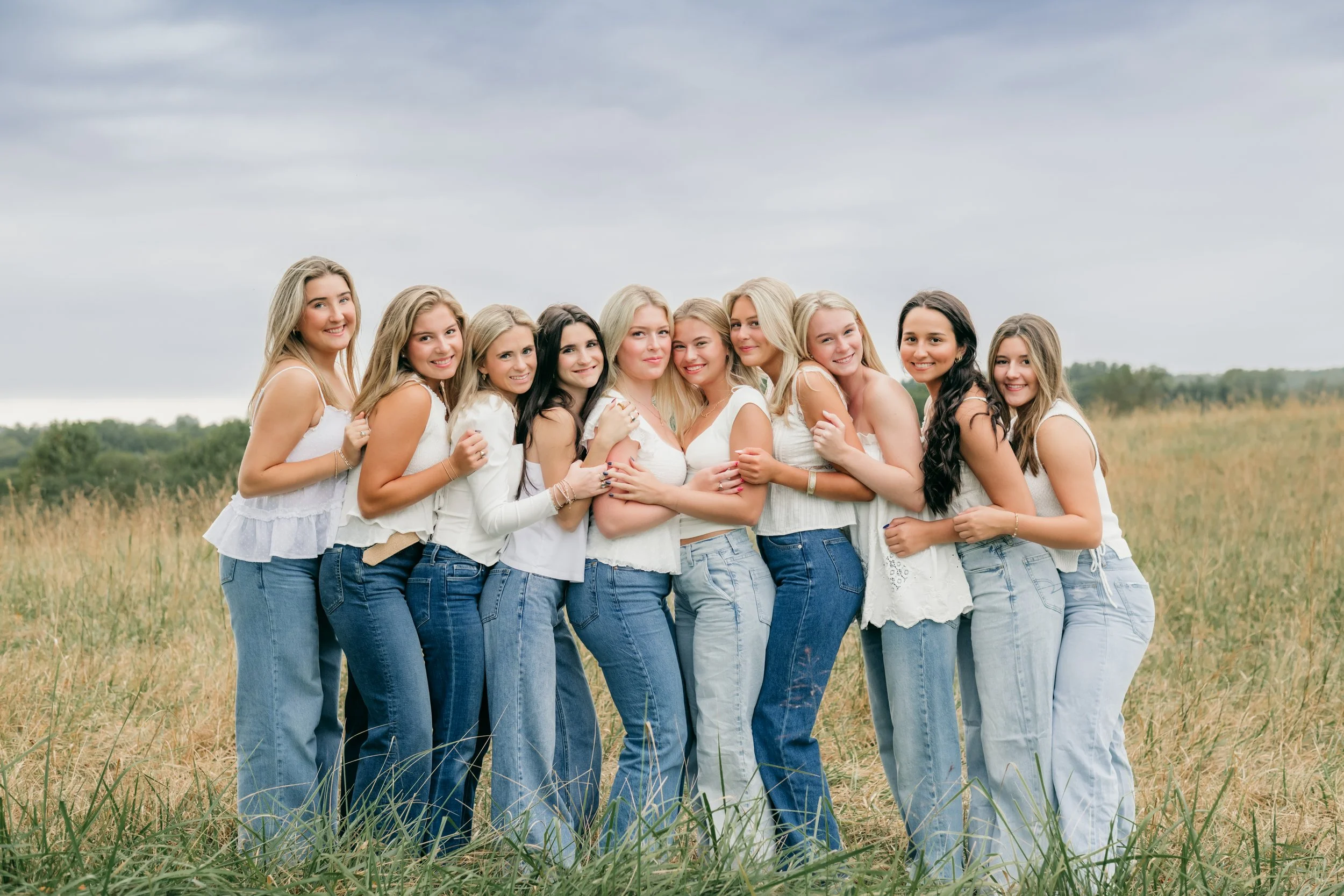 A group of thirteen young women standing together outdoors in a grassy field on a cloudy day, all wearing white tops and blue jeans, smiling and hugging each other.