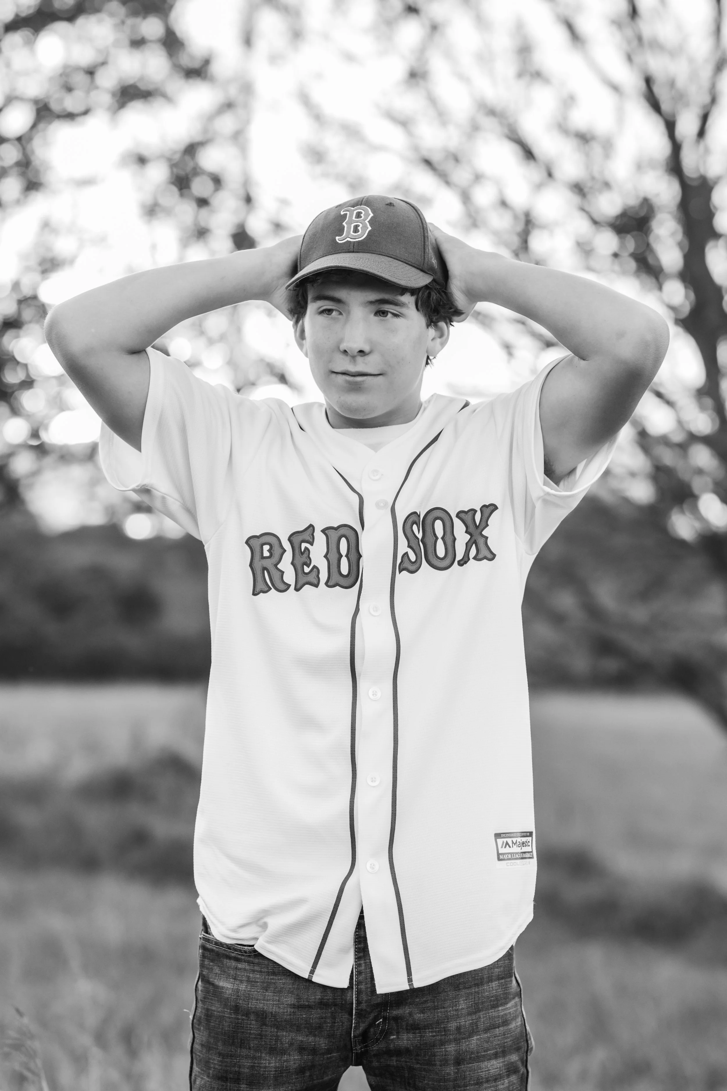 A young male teenager wearing a Boston Red Sox baseball jersey and cap, standing outdoors with trees in the background, holding his hands behind his head.