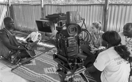 A woman is being interviewed or recorded on video by a team of three people in a room with corrugated metal walls. They are sitting on a patterned rug, and a professional video camera is positioned in front of them.