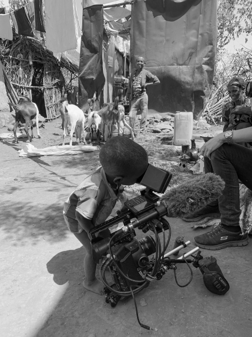 A young boy looking through a professional video camera in a rural outdoor setting, with goats and a man standing in the background.