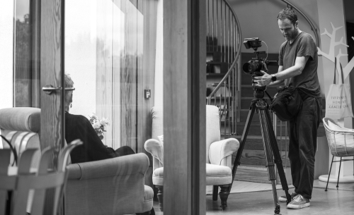 A man operating a video camera on a tripod, filming a woman sitting in a cozy indoor space with large windows and furniture.