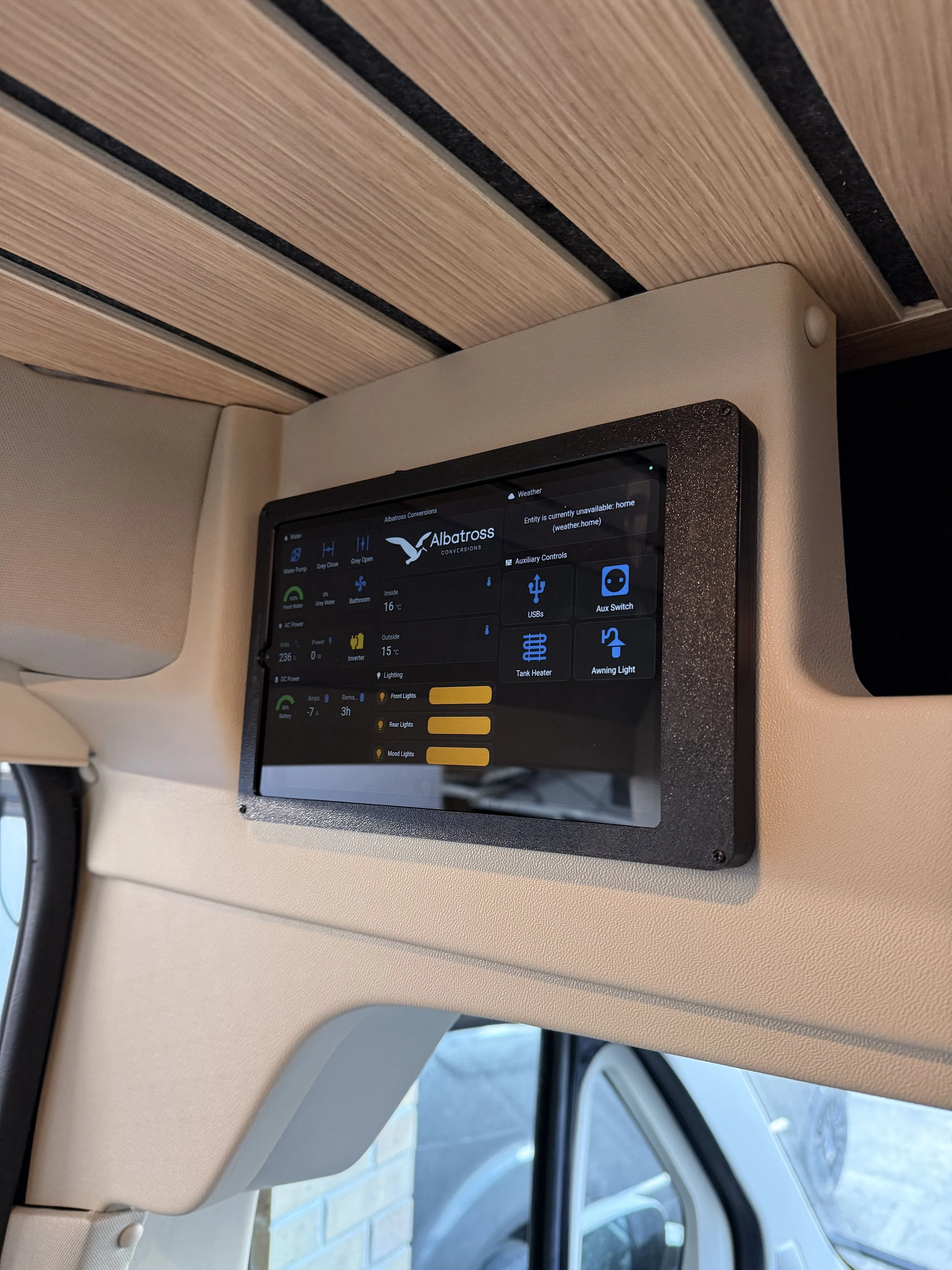 Digital control panel mounted on a light-colored wall in a modern kitchen. The screen displays various settings and information related to water, lighting, AC power, and battery status.