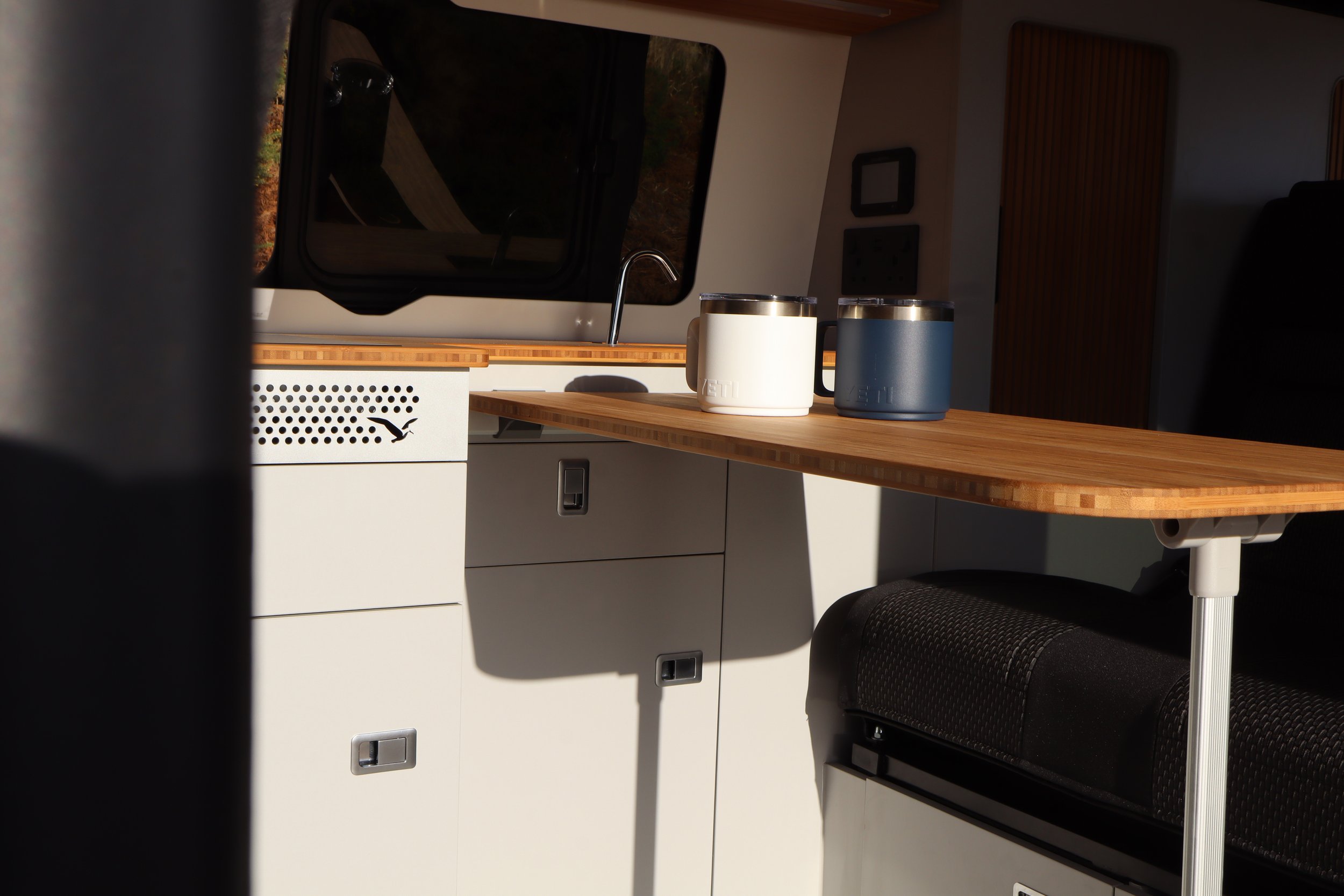 Interior of a camper van with a wooden countertop, two mugs, a window, and cabinet storage.