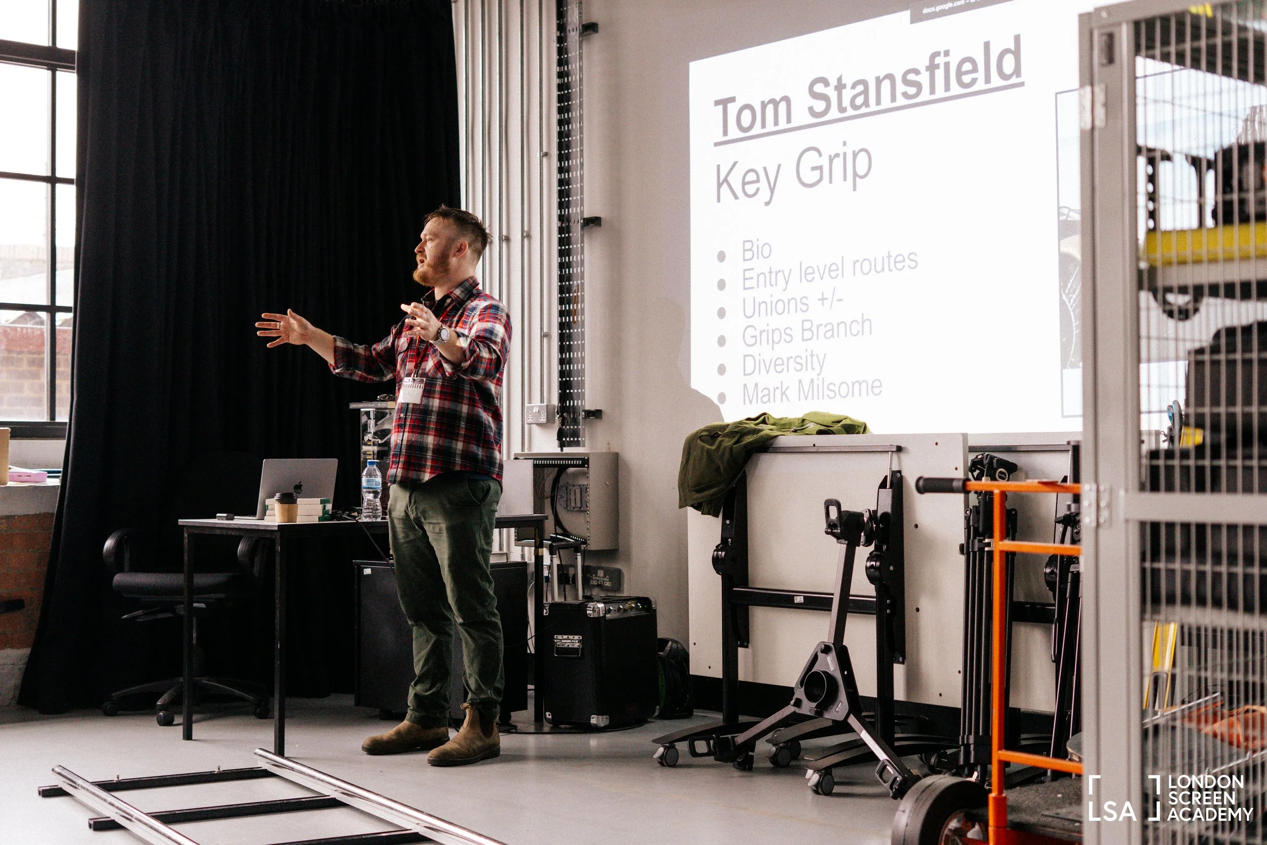A man giving a presentation in a conference room next to a large screen display with a PowerPoint slide. The slide shows the name 'Tom Stansfield' and the title 'Key Grip' along with bullet points. The room has large windows, a black curtain, and var