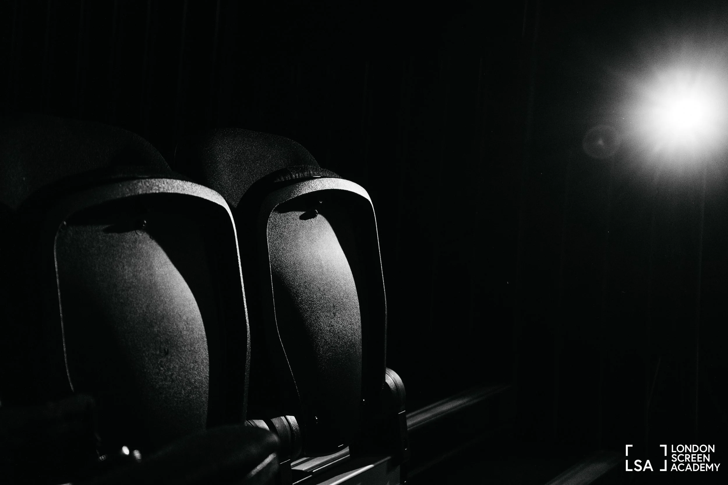 Black and white photograph of empty movie theater seats with a bright light in the distance in a dark room.