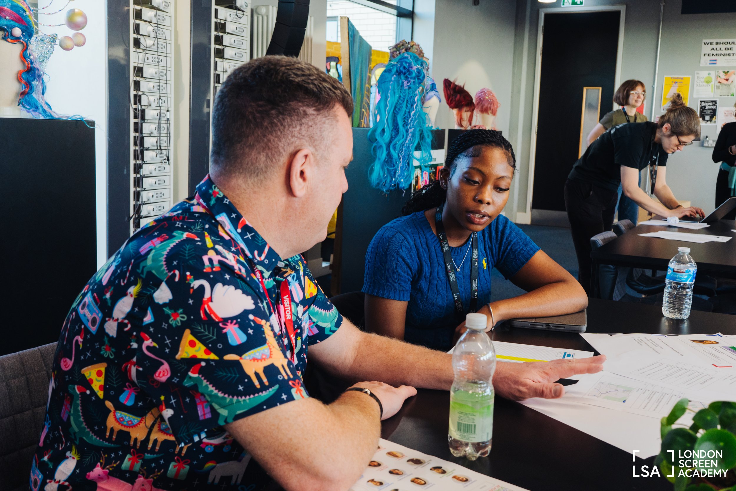 Two people sitting at a table in a creative workspace, discussing documents. One wears a colorful holiday-themed shirt, and the other wears a blue top. In the background, other individuals work at tables, with colorful wigs and hairpieces displayed o