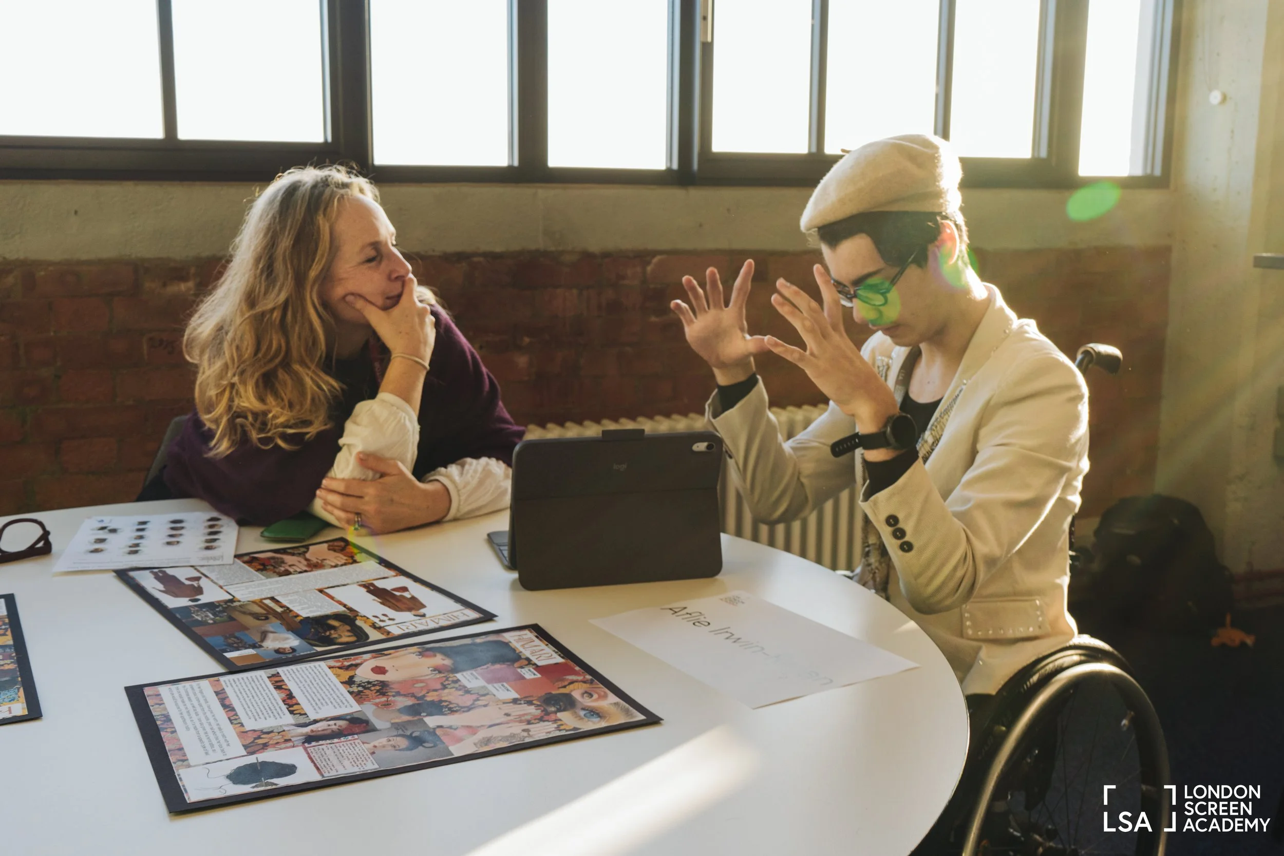 A young woman in a wheelchair and a white blazer engages in a conversation with an older woman with long curly blonde hair, seated at a table with various magazines and photos in a room with large windows and sunlight.