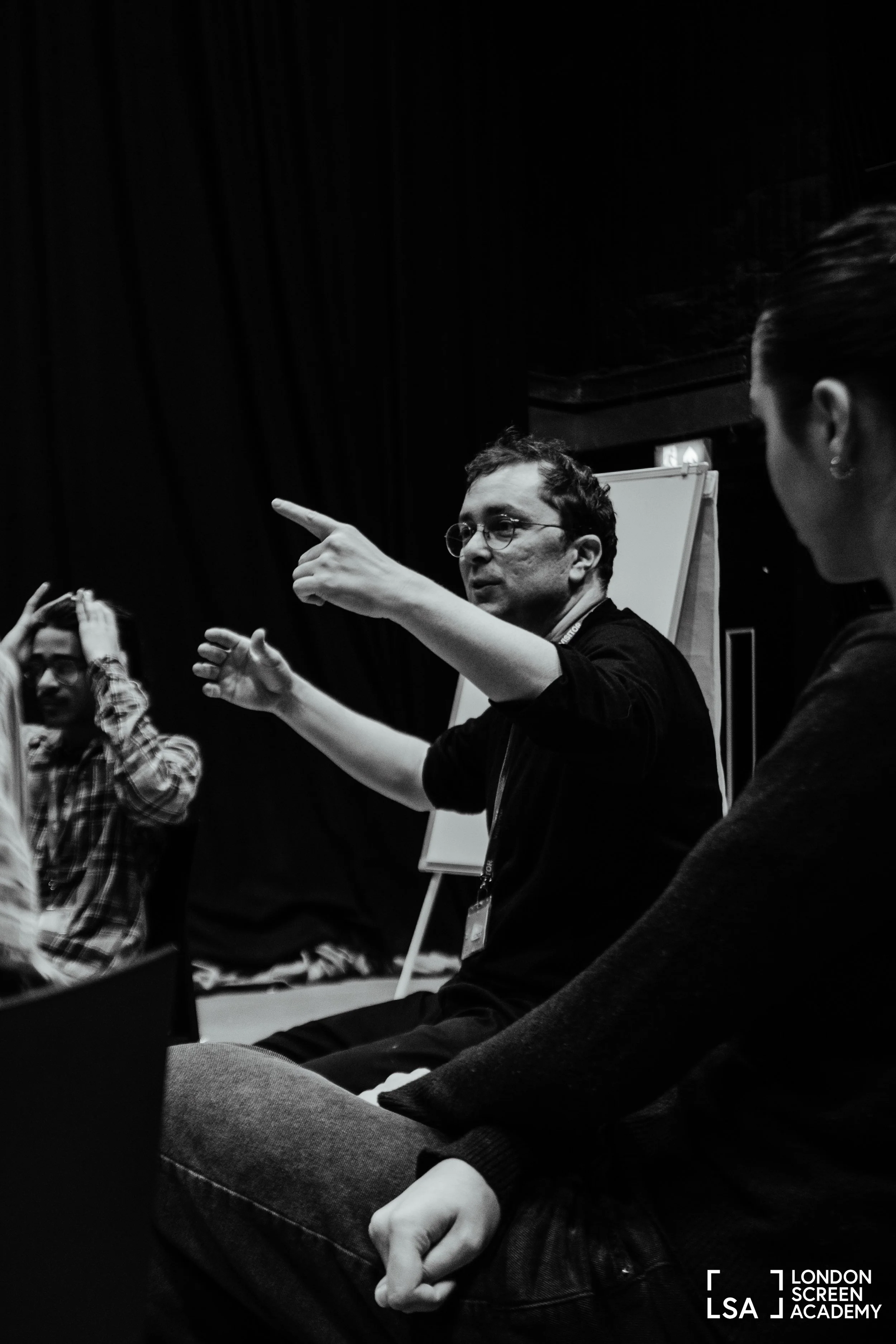 A man with glasses and curly hair gesturing with his hands while speaking during a discussion at London Screen Academy, with others seated nearby. The setting appears to be an indoor workshop or seminar.