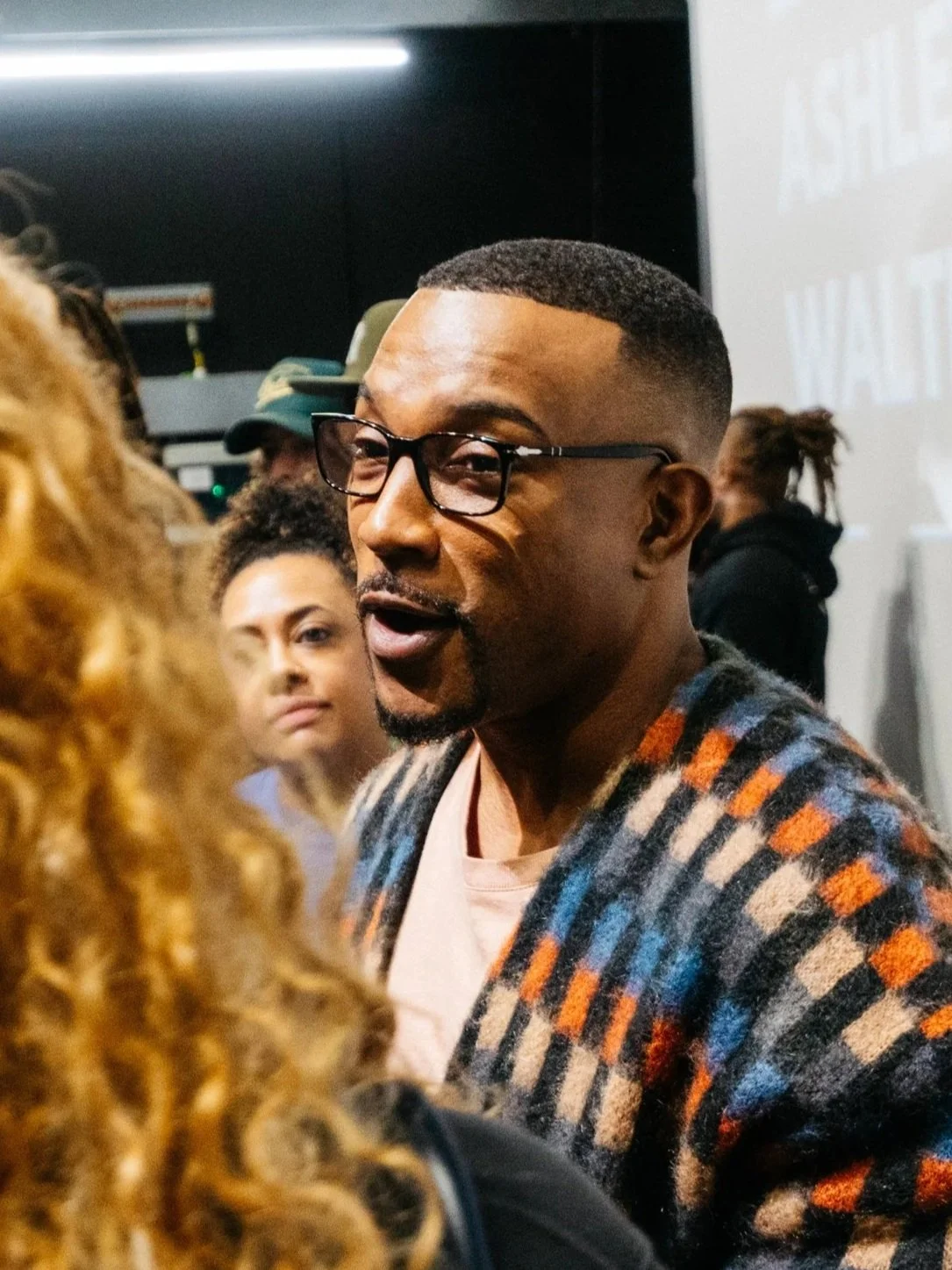 Actor Ashley Walters  wearing glasses and a plaid jacket is talking, with two women and a man in the background, possibly at a private masterclass.