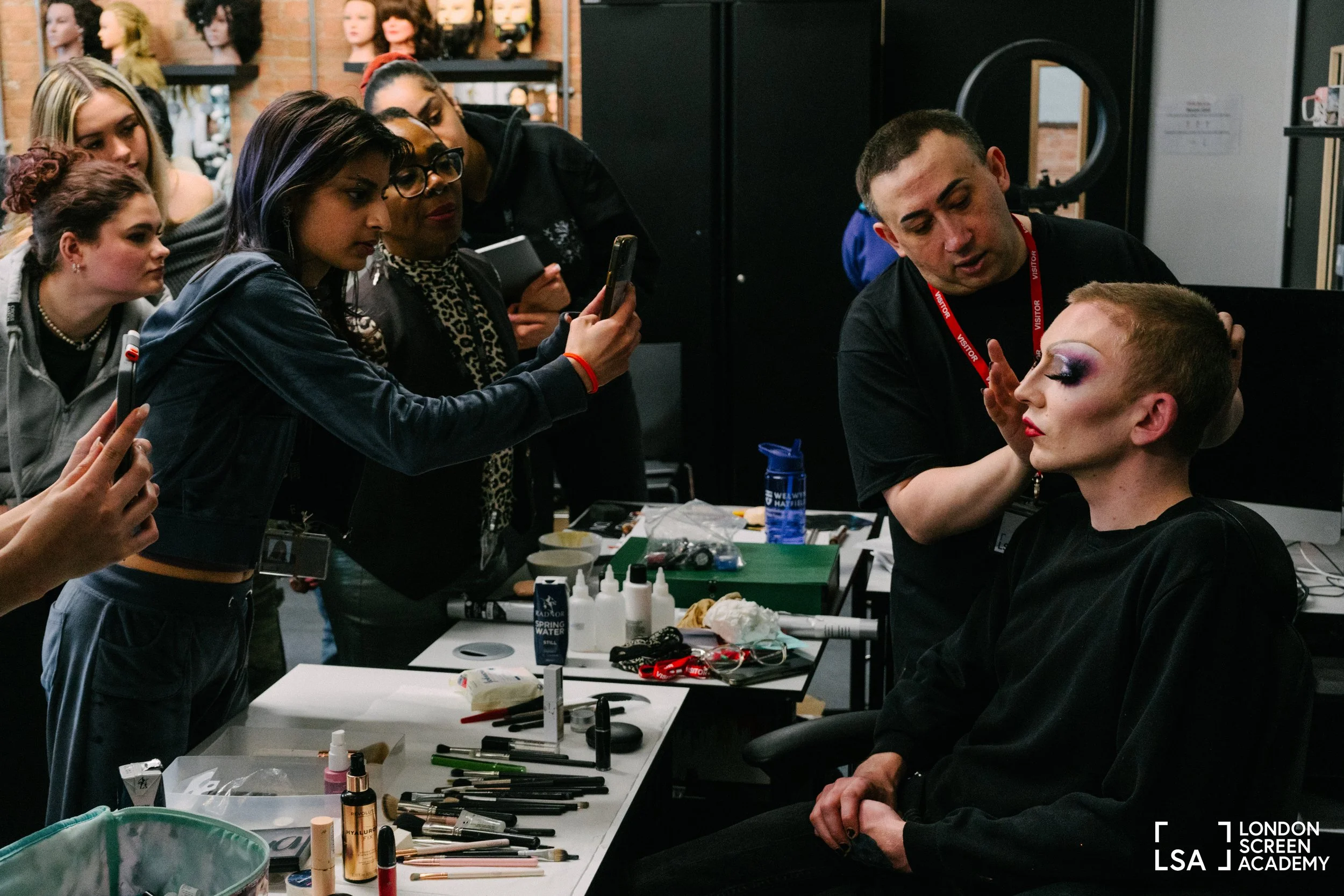 Makeup artists and models at a makeup studio, with one model having elaborate makeup, while makeup tools and supplies are on the table.