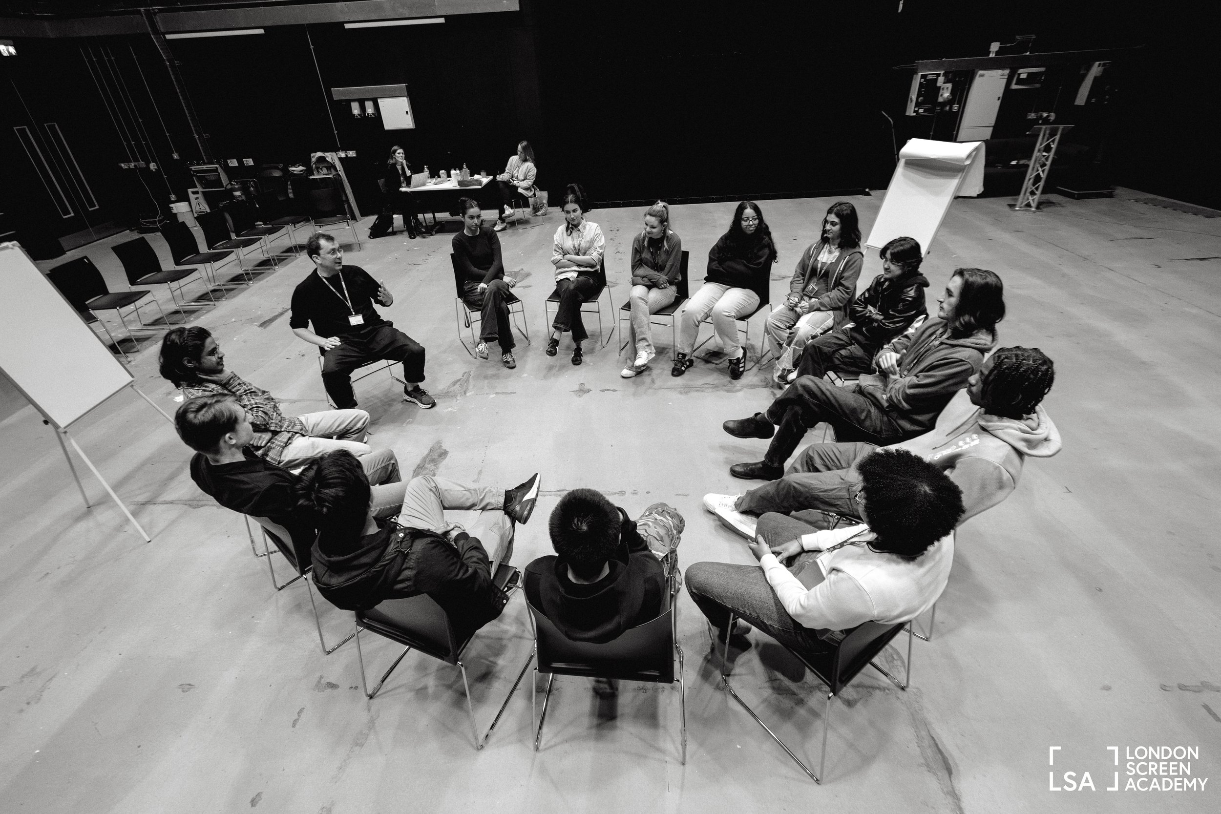 A black-and-white photo of a group of people sitting in a circle in a large indoor space, participating in a discussion or workshop, with some individuals taking notes while others listen.