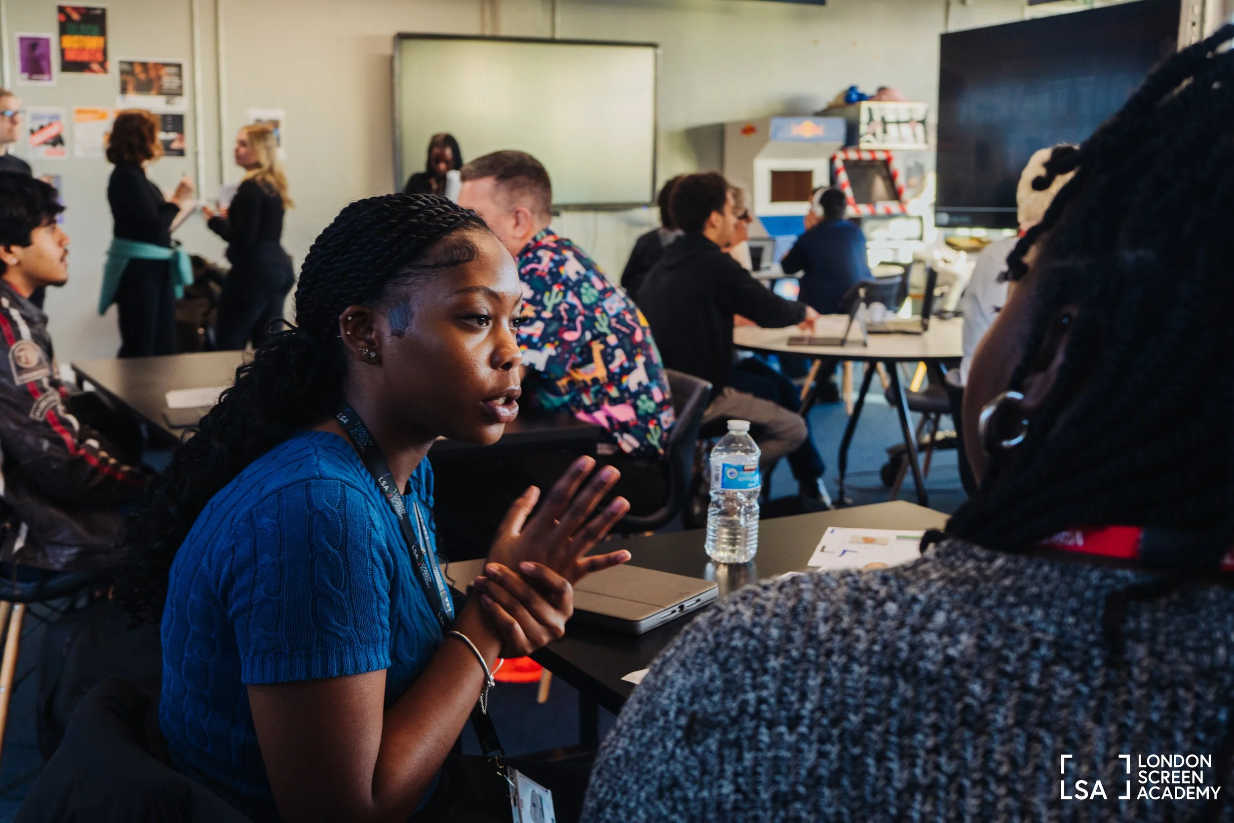 A young woman with braided hair and a blue shirt is speaking and gesturing with her hands to another person seated across from her in a classroom or conference room. Several other people are sitting at tables, some engaging in conversations, with a w