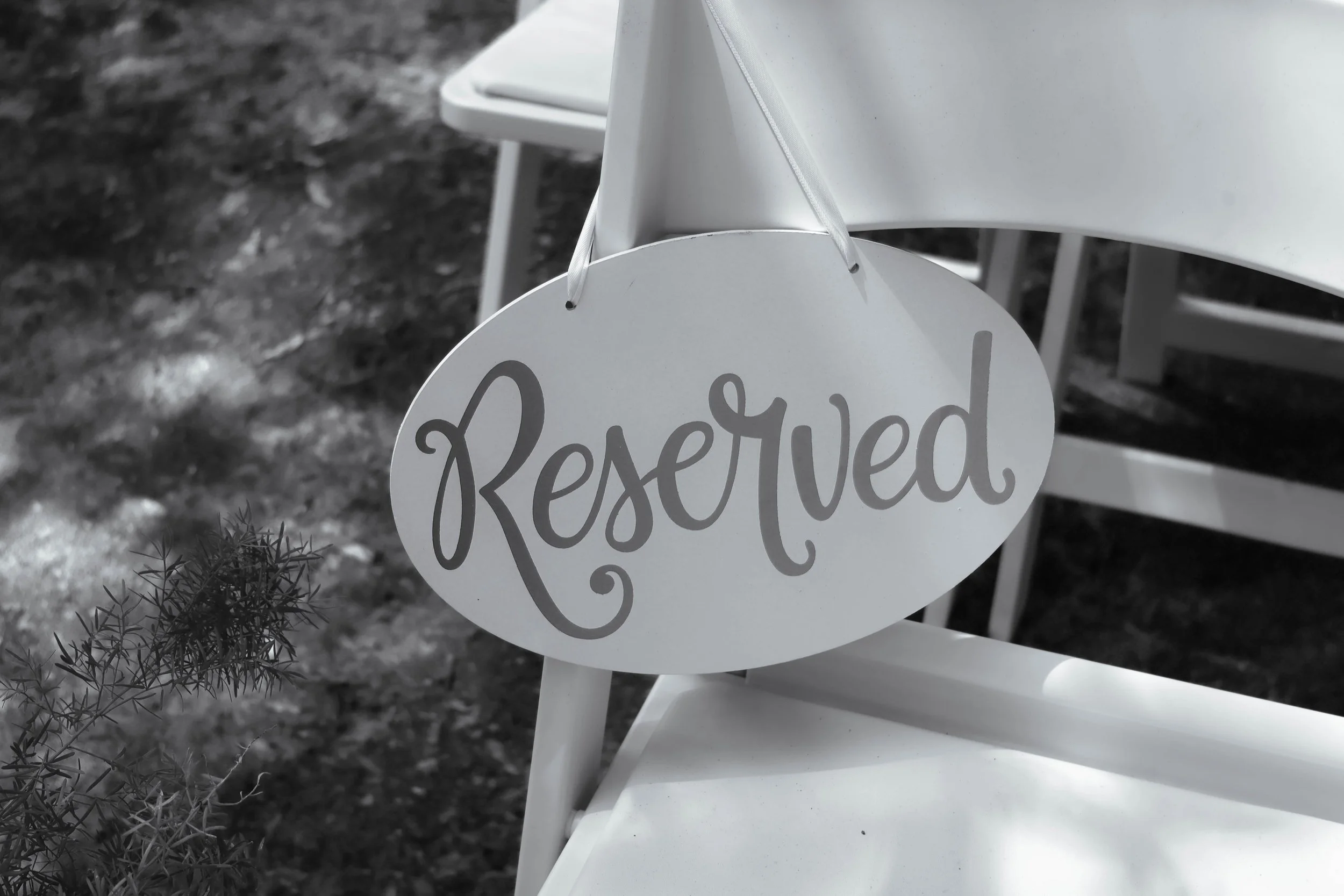 A reserved sign hanging from a white chair in an outdoor setting with dirt and a small bush nearby.