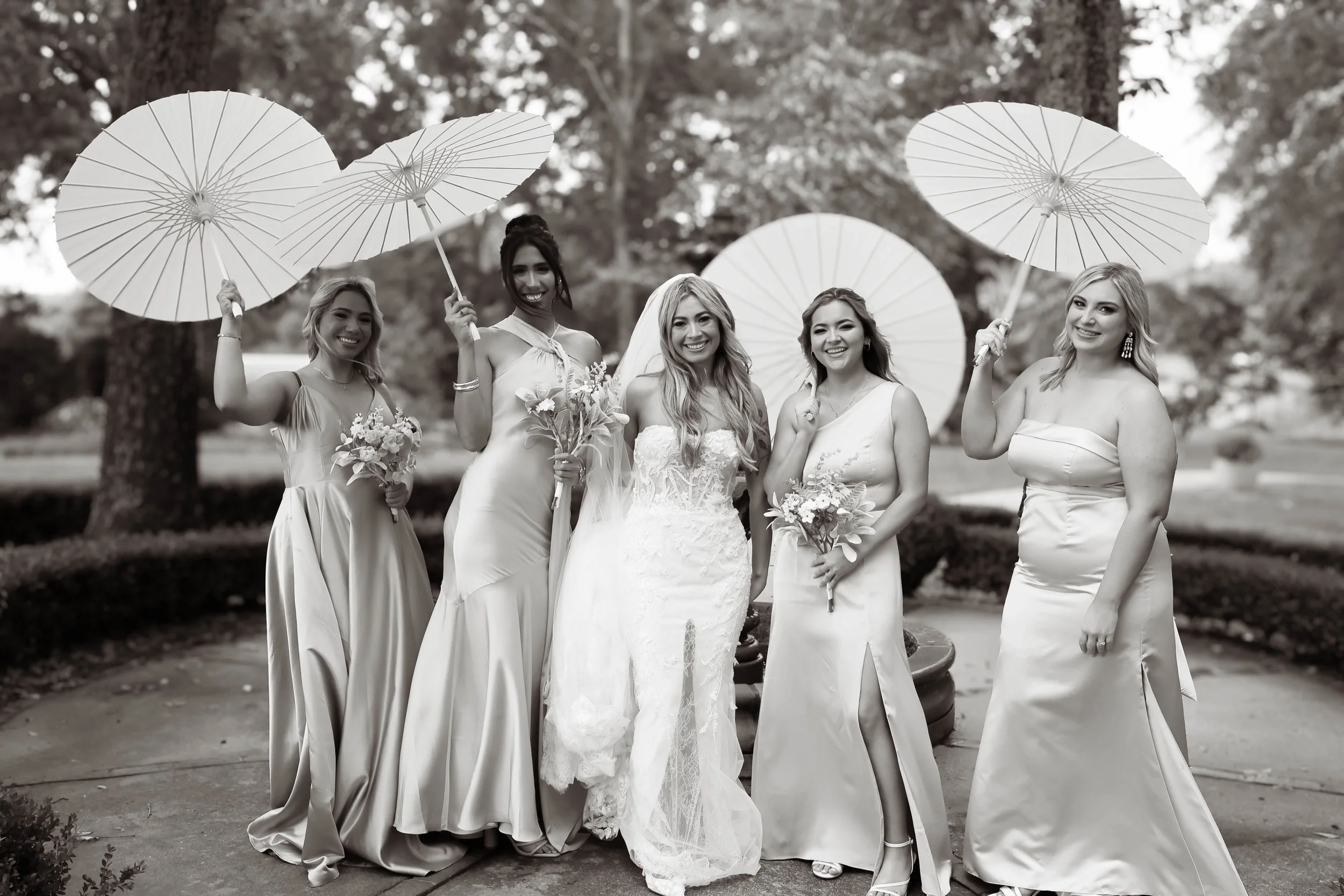 A group of six women, including a bride in a wedding dress, standing outdoors with trees in the background. They are smiling and holding parasols and bouquets, celebrating a wedding.