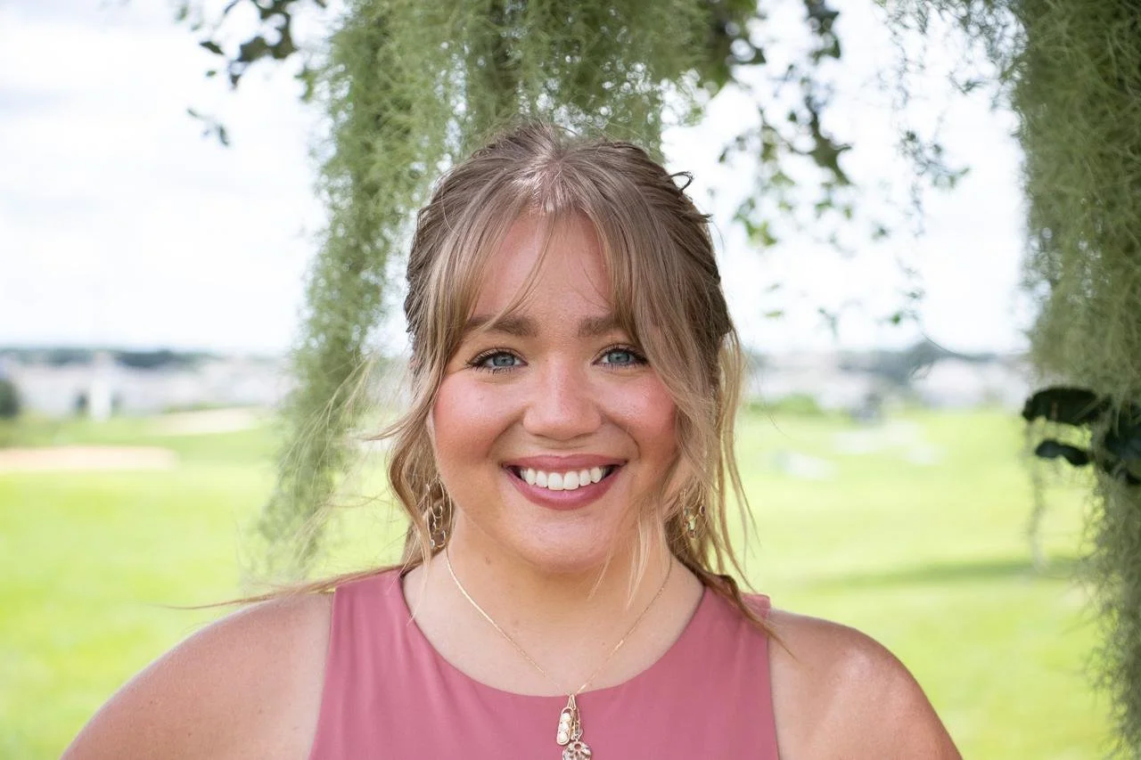 Smiling woman in a pink dress outdoors with greenery in the background.
