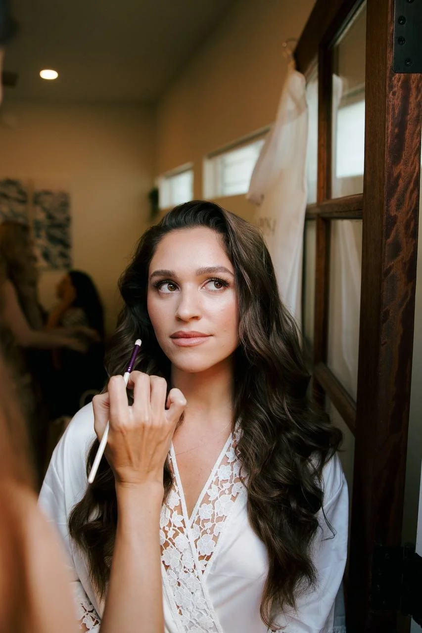 A woman with long, dark wavy hair and light skin, wearing a white strapless top and a delicate gold necklace, standing in an indoor setting with neutral-colored walls and mirrors in the background.