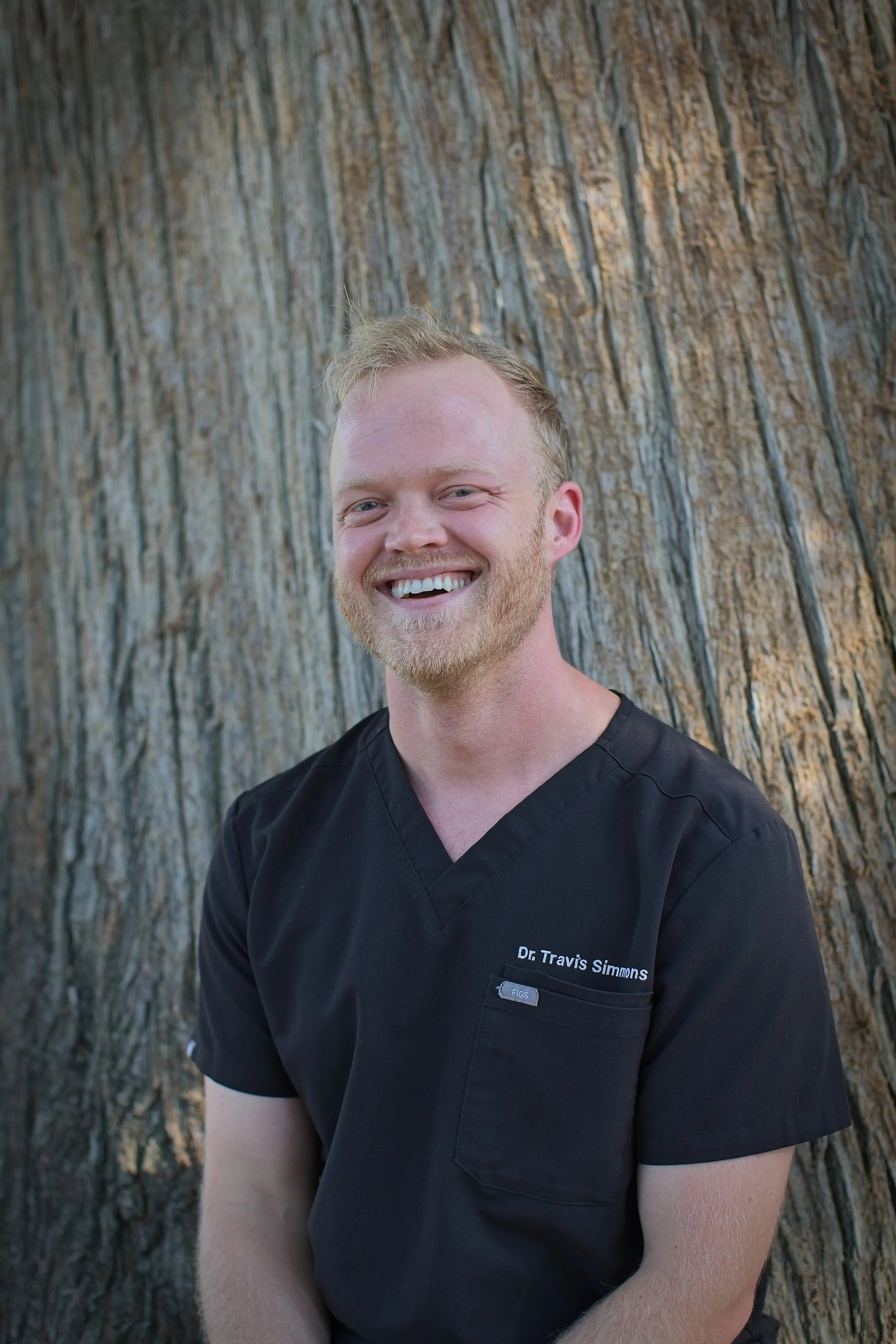 A young man with red hair and a beard, wearing a light gray blazer and light blue shirt, smiling outdoors with trees and greenery in the background.